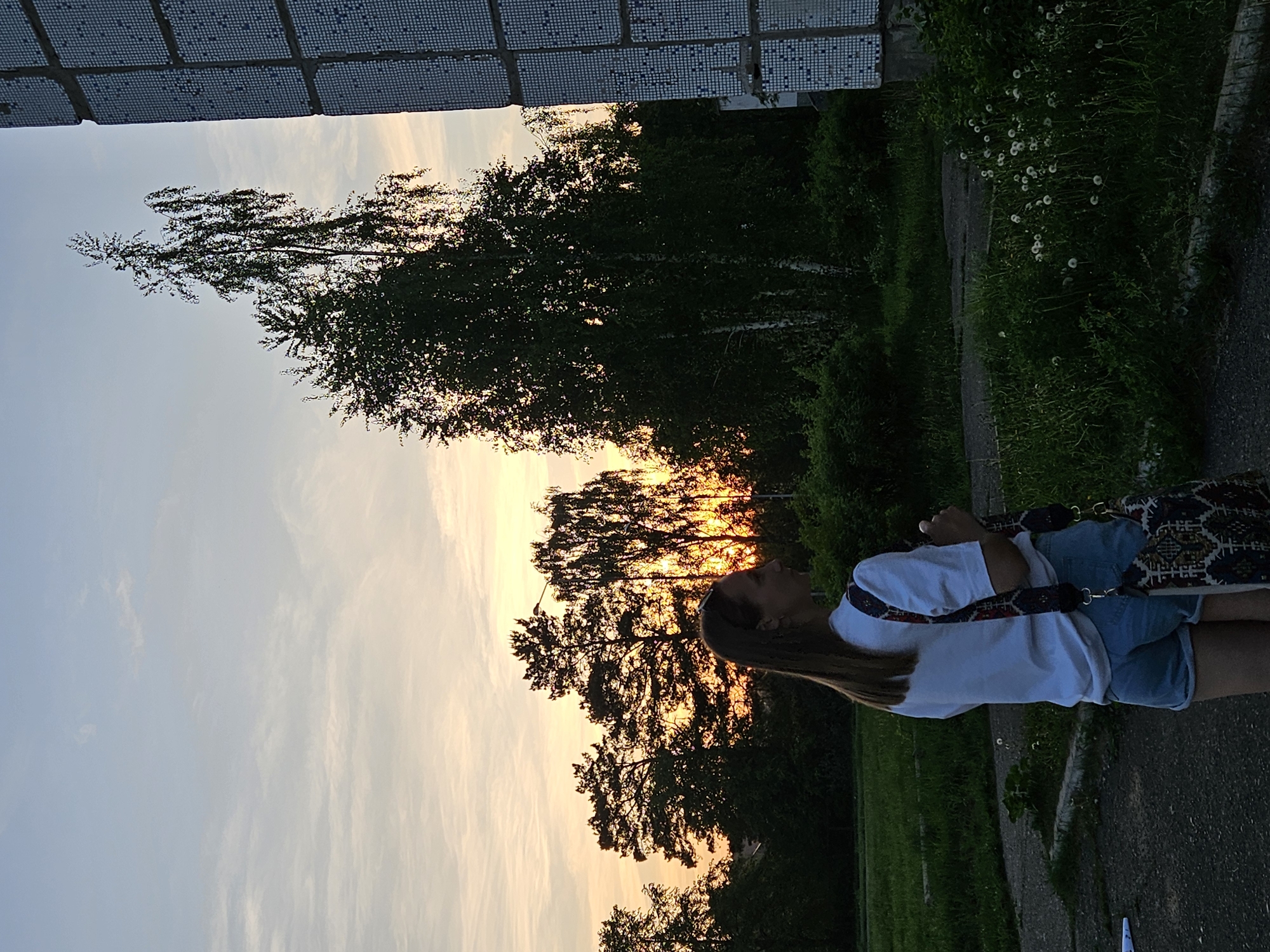 The image depicts a woman standing on a sidewalk in the city of Chaikovsky, Russia. She is wearing a white t-shirt, denim shorts, and a patterned handbag. Behind her are trees silhouetted against a sunset sky. To her right is a grassy area with weeds and dandelion seed heads. A tiled building overhangs the top of the image.