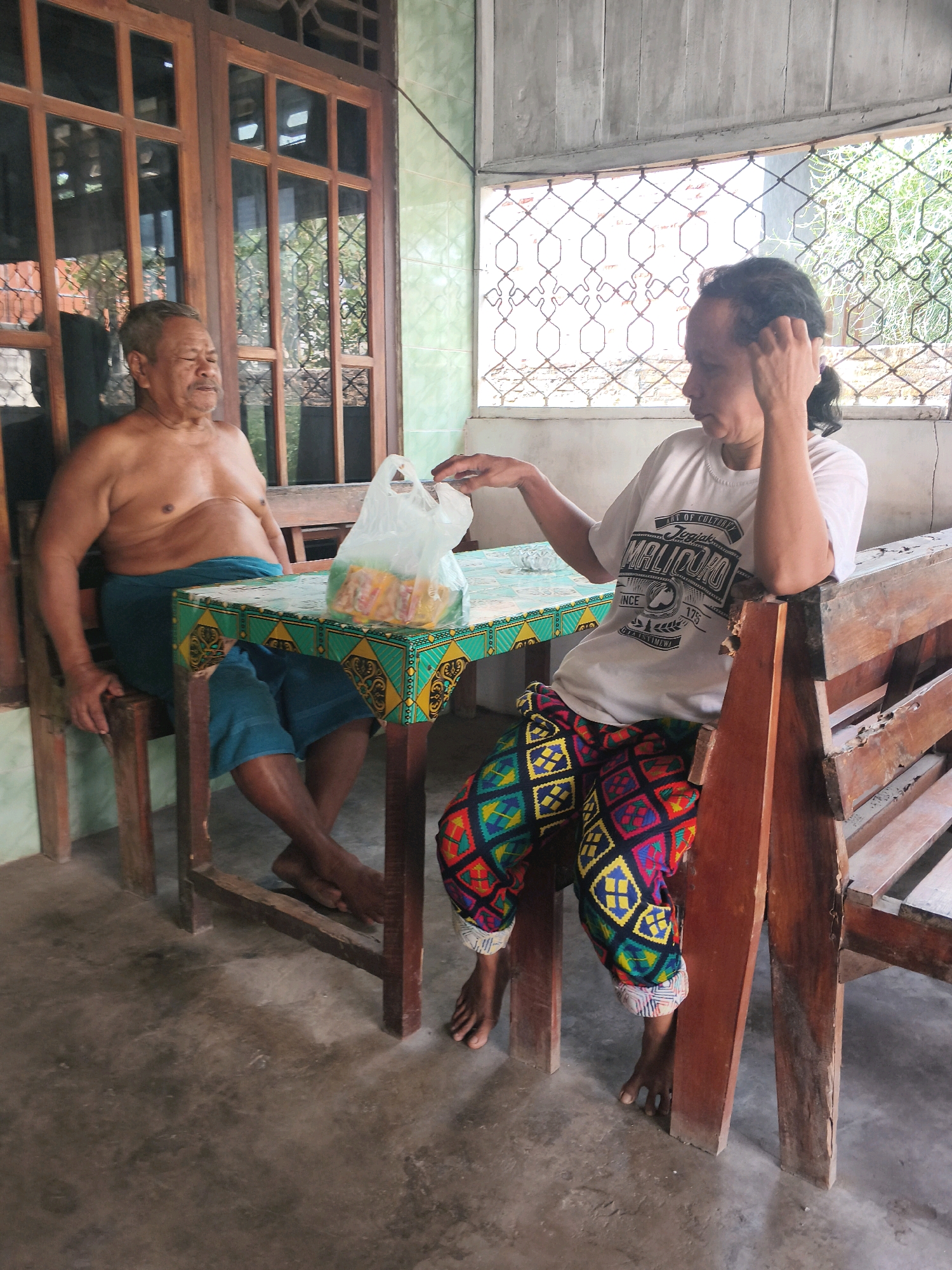 Two individuals are seated at a table outdoors, likely on a porch, in Way Galih, Indonesia. The individual on the left, a shirtless man with tan skin and graying hair, wears a blue sarong or wrap. He is seated on a wooden chair, leaning back slightly with his legs crossed. The individual on the right, with darker skin and dark hair pulled back, wears a white t-shirt and vibrantly patterned pants. They are seated on a wooden bench and are leaning forward, with their right hand touching their hair and their left hand resting on the table.
On the table, which has a green, yellow, and black patterned cloth, is a clear plastic bag containing what appears to be food items, possibly snacks. The environment suggests a tropical climate; there are large wooden-framed windows with glass panes and a screened opening, allowing a view of lush green foliage outside. The floor is concrete. The wooden furniture, including chairs and a bench, shows signs of wear. The overall impression is a casual, everyday scene in a residential outdoor area.