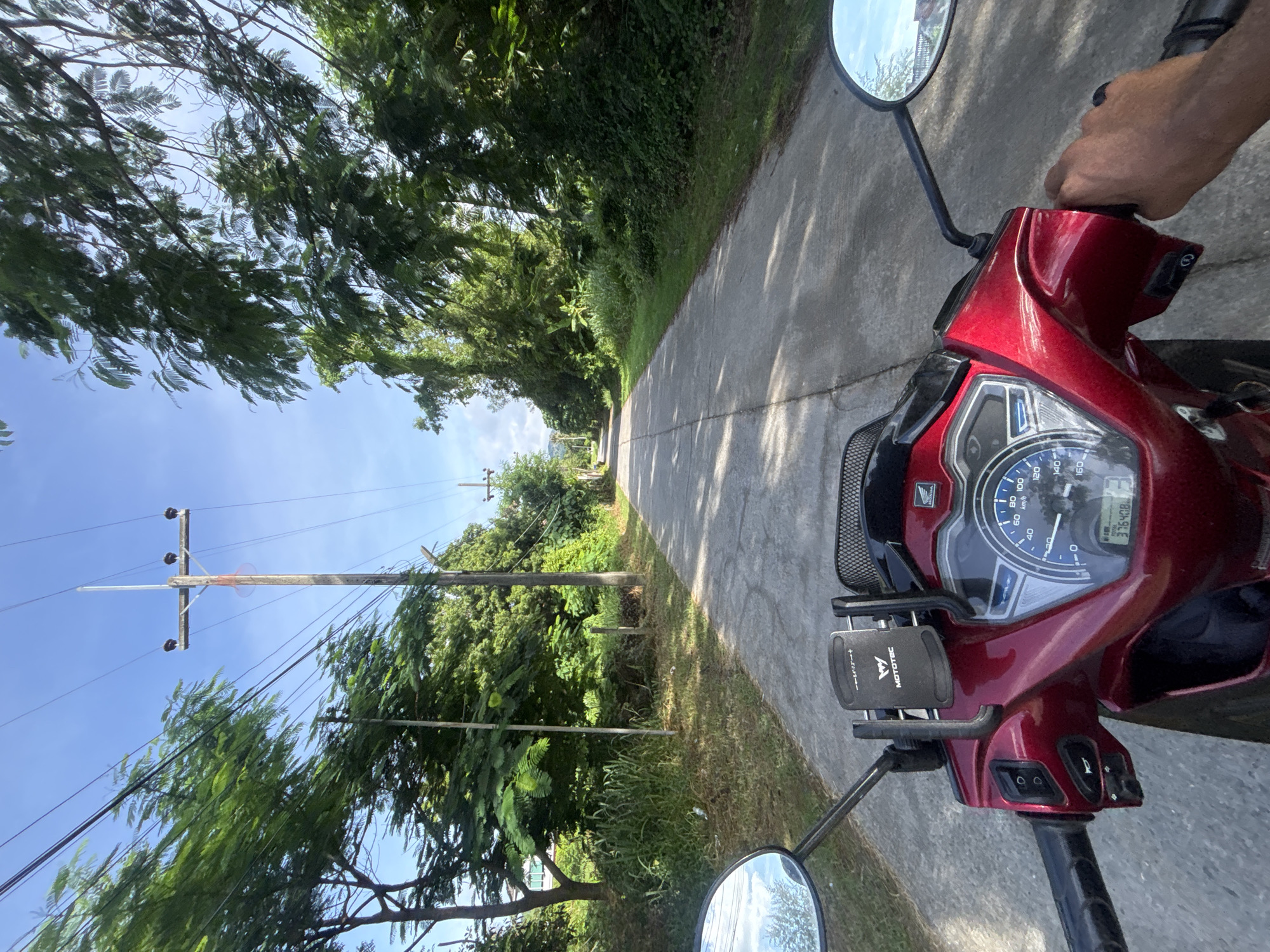 The image depicts a first-person perspective of someone riding a red scooter on a paved road in Chalong, Thailand. The road is lined with lush green trees and vegetation, with a clear blue sky overhead. Utility poles with power lines run along the side of the road. The scooter's dashboard and handlebars are visible in the foreground, including a phone mount with the brand "MOTOTEC" clearly displayed. The rider's hand is visible on the right handlebar. The scene suggests a sunny day in a tropical or subtropical environment, likely a rural or suburban area of Chalong, Thailand, given the presence of dense greenery and utility infrastructure.