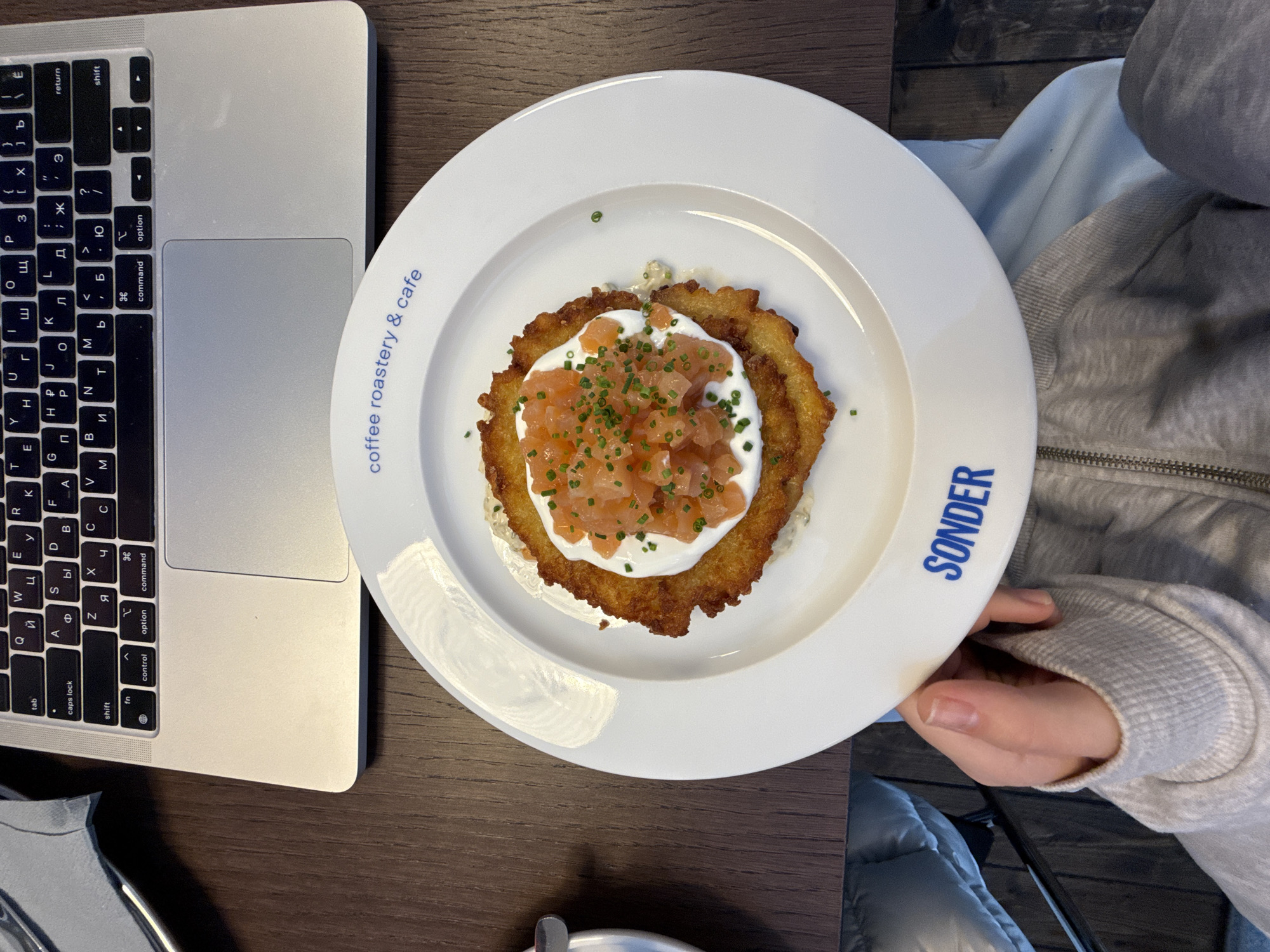 The scene presents a top-down view of a table setting, likely in a cafe or restaurant. A laptop is positioned on the left side of the frame, its keyboard partially visible with Cyrillic characters interspersed among the standard QWERTY layout. To the right of the laptop, a white plate dominates the center of the image. The plate holds a potato pancake, topped with a layer of cream and diced salmon, garnished with green chives. The words "coffee roastery & cafe" are printed around the edge of the plate, along with the name "SONDER" in blue. To the right of the plate, a person's arm and hand are visible. The person is wearing a light gray sweatshirt or jacket. The table surface is dark brown and textured. The scene may be in Belgrade, Serbia due to the Cyrillic keyboard.