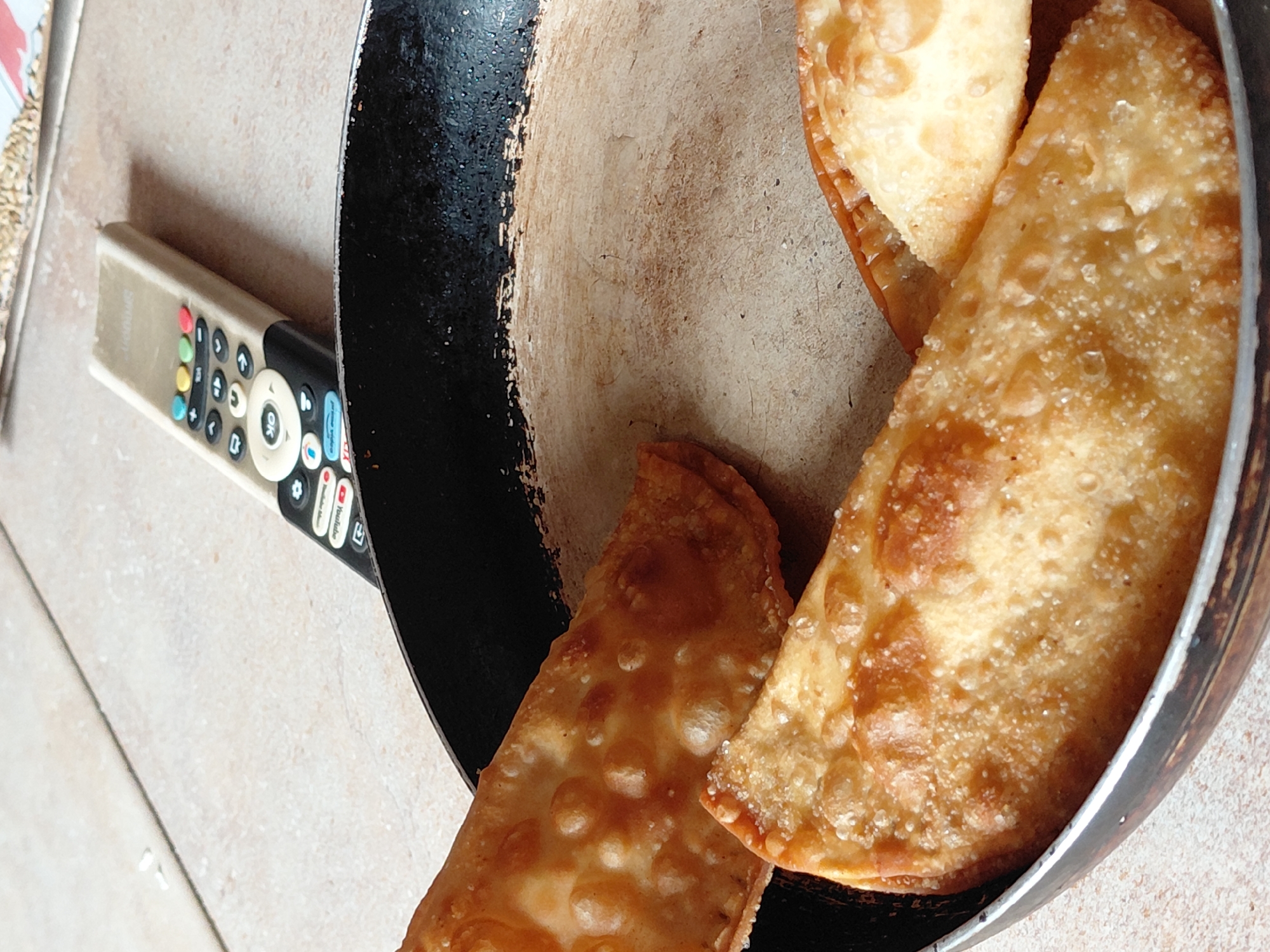 A close-up, high-angle shot captures several golden-brown, deep-fried pastries resting inside a black frying pan, positioned on a light-colored tiled surface. The pastries, resembling *pasteles* or *empanadas*, have a crisp, bubbly texture, and some appear to be sprinkled with fine sugar crystals. The frying pan shows signs of use, with wear on its interior coating.
Adjacent to the pan, a beige remote control with a black top section lies on the tiles. The remote features a variety of buttons, including prominent colorful ones (red, green, yellow, blue) and numerous gray and white buttons. Text "SMART" is faintly visible on its body. Key buttons visible include "OK" in the center, a gear icon (likely for settings), a "YouTube" logo, and what appears to be a home icon.
The scene depicts a casual, domestic setting, likely indoors in Santa Ana, Venezuela. The pastries appear freshly cooked, suggesting they are either cooling or awaiting consumption. The diffused lighting indicates it is likely daytime. No people are visible, and no specific activity beyond the presence of the food and remote control is taking place.
