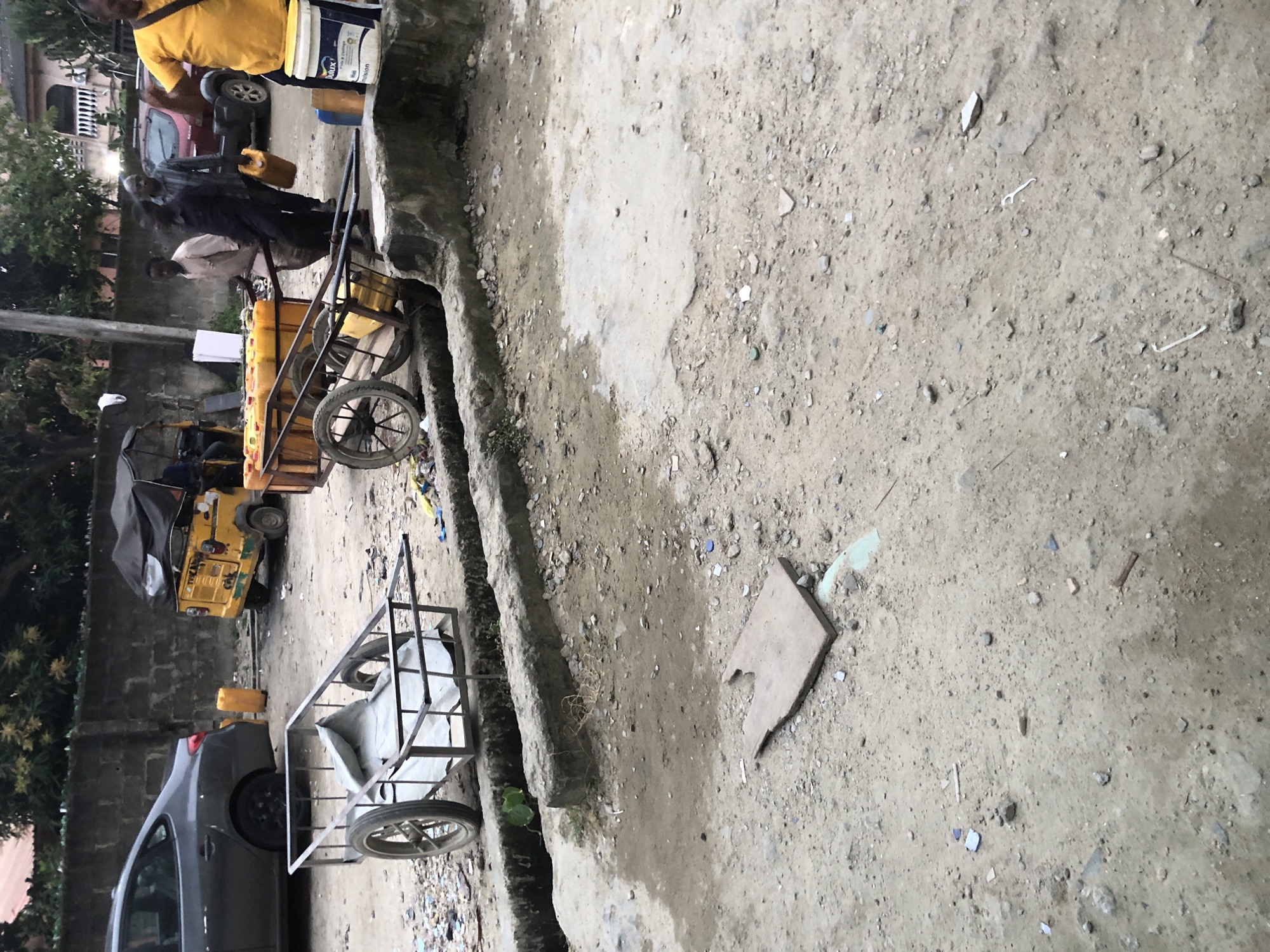 In Ado, Nigeria, an outdoor scene captures a dusty, unpaved area with some debris scattered across the ground. In the mid-ground, a two-wheeled cart with a metal frame and a grey tarp is positioned. Behind this cart, another larger cart is loaded with multiple yellow jerrycans, some of which have red caps. Three men are visible near this cart. One man, wearing a patterned shirt, is holding a yellow jerrycan and appears to be speaking to another man in a dark shirt who is pushing the cart. A third man, in a light-colored outfit, stands to the left. Further back, a yellow motorized tricycle with a black canopy is parked, and a dark-colored sedan is visible to its left. A person wearing a bright yellow t-shirt stands to the far right, partially obscuring a white bucket with a blue and red logo. A concrete barrier runs along the foreground, separating the street from a lower-lying area with some vegetation.