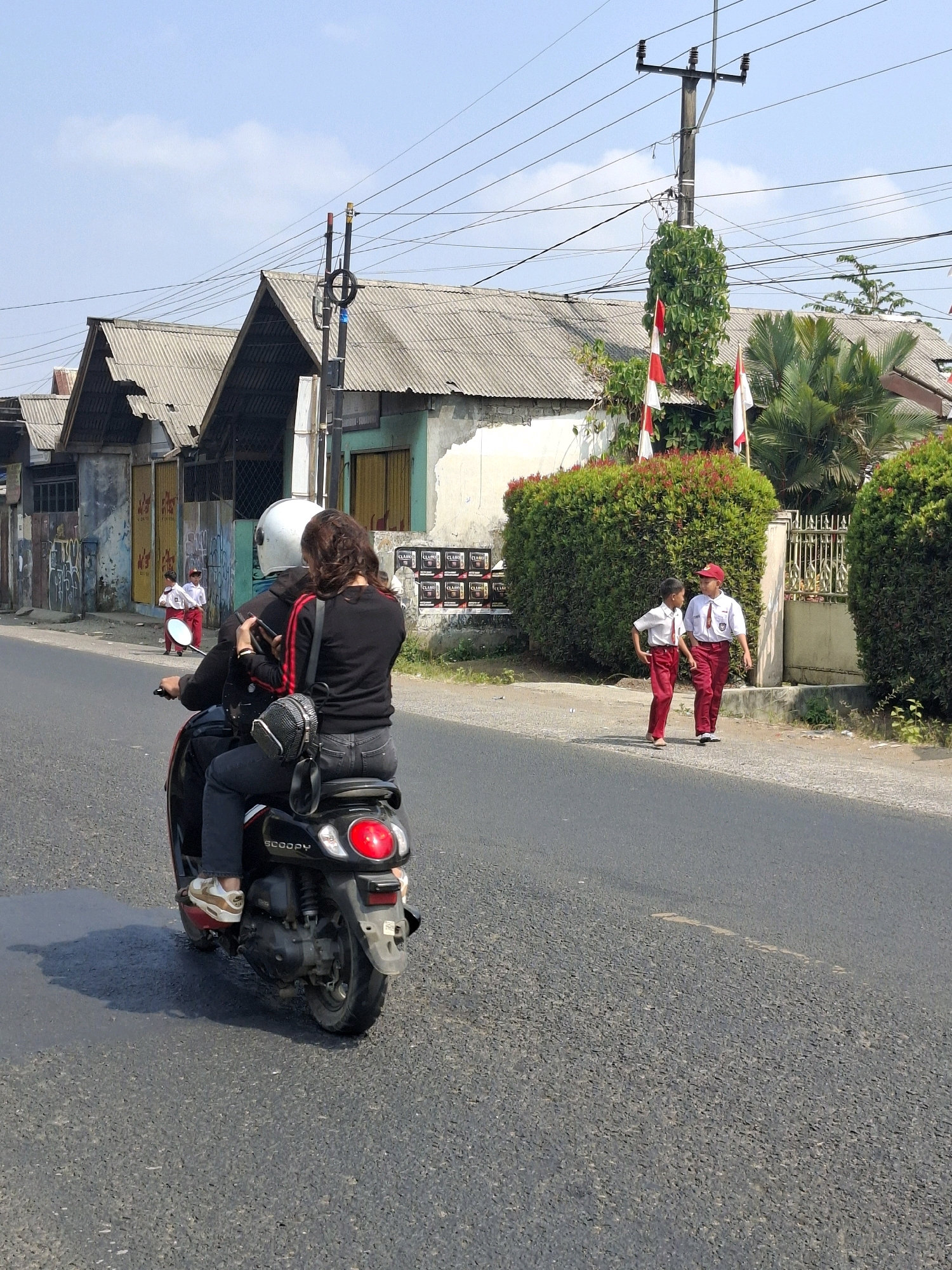 The image depicts a street scene in Lembursawah, Indonesia, under a clear sky. In the foreground, two individuals are riding on a black scooter. The person in the back, identified as a woman, is wearing a black long-sleeved shirt with red stripes on the sleeves, black jeans, and is holding a smartphone. She has a black crossbody bag with a patterned design. The person in front, wearing a white helmet, is operating the scooter. The scooter is a Honda Scoopy.
On the right side of the street, two young boys in school uniforms, consisting of white shirts and red pants, are walking side-by-side. They appear to be engaged in conversation. Behind them, a manicured hedge runs along the sidewalk, and a white fence can be seen. Several Indonesian flags are displayed on flagpoles, some attached to a utility pole and others near the hedge.
The background features a row of modest buildings with corrugated metal roofs. Overhead, a network of electrical wires crisscrosses the sky, supported by utility poles. The road is paved asphalt, and a shadow of the scooter is visible on the left side of the road. The overall environment suggests a residential or semi-commercial area in a tropical climate.