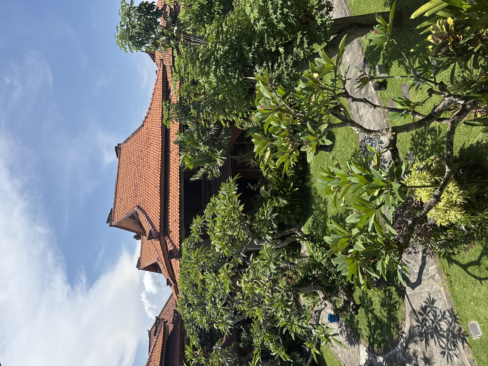 This image shows a view of a building with a red tile roof in Unknown, Indonesia. The sky is blue with some clouds. Green trees and plants surround the building. A concrete pathway runs through the green space.