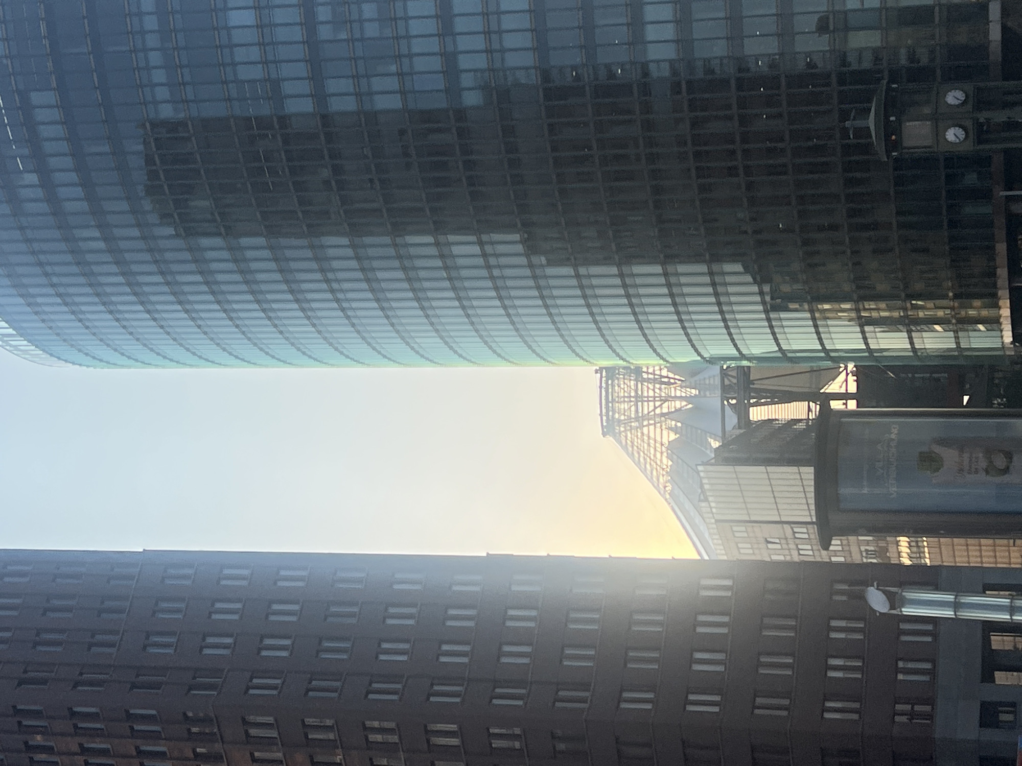 The image shows a view of the Potsdamer Platz in Berlin, Germany. The scene features two prominent modern skyscrapers, one a dark, rectangular building on the left and a curved, glass-facade building on the right. Between them, in the background, is a distinctive conical, glass-covered structure, which is part of the Sony Center. The sky is bright and hazy, suggesting it is either sunrise or sunset, with the light reflecting off the buildings. In the foreground, on the right side, a traditional Berlin column clock with two faces is visible. The overall impression is one of a modern urban landscape.