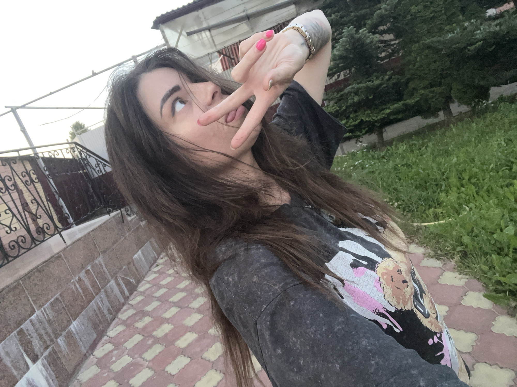 A young woman with long, dark brown hair is taking a selfie outdoors. She is wearing a dark t-shirt with a graphic print. Her left hand is raised, with her index and middle fingers extended in a peace sign, and her tongue is sticking out. She is looking up and to her left. Behind her, a paved walkway with decorative tiles leads to a stone staircase with a black metal railing. Further back, there is green grass, trees, and a building with a tiled roof. The scene appears to be a residential area in the city of Krasnoe-na-Volge, Russia.