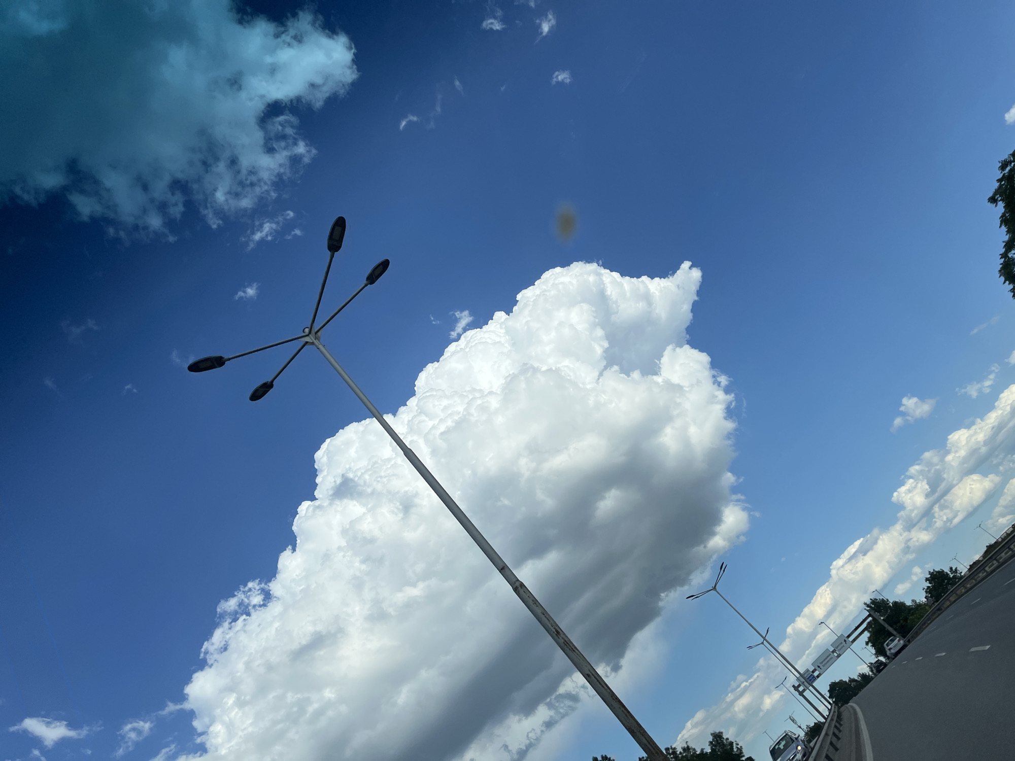 A low-angle shot of a bright blue sky with fluffy white clouds and a tall lamppost with multiple lights. The lamppost is positioned to the left of the frame, extending diagonally upwards. A highway with cars driving on it is visible in the lower left portion of the image, with trees lining the side of the road. The image is taken from a moving vehicle, as suggested by the road and its perspective. The overall scene is one of a sunny day on a highway.