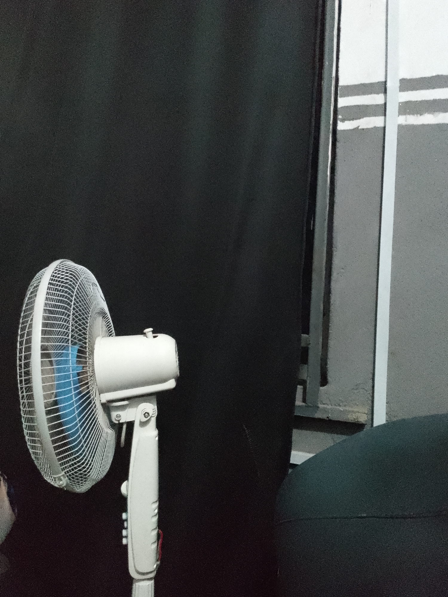 A white oscillating fan with a blue fan blade is positioned on the left side of the frame, set against a dark background. To the right of the fan, a wall is visible with horizontal grey stripes. In the foreground, a dark, textured object, possibly furniture, occupies the bottom right corner of the image. The lighting is dim, with some light reflecting off the fan's metal grille and the wall. The overall scene appears to be indoors.