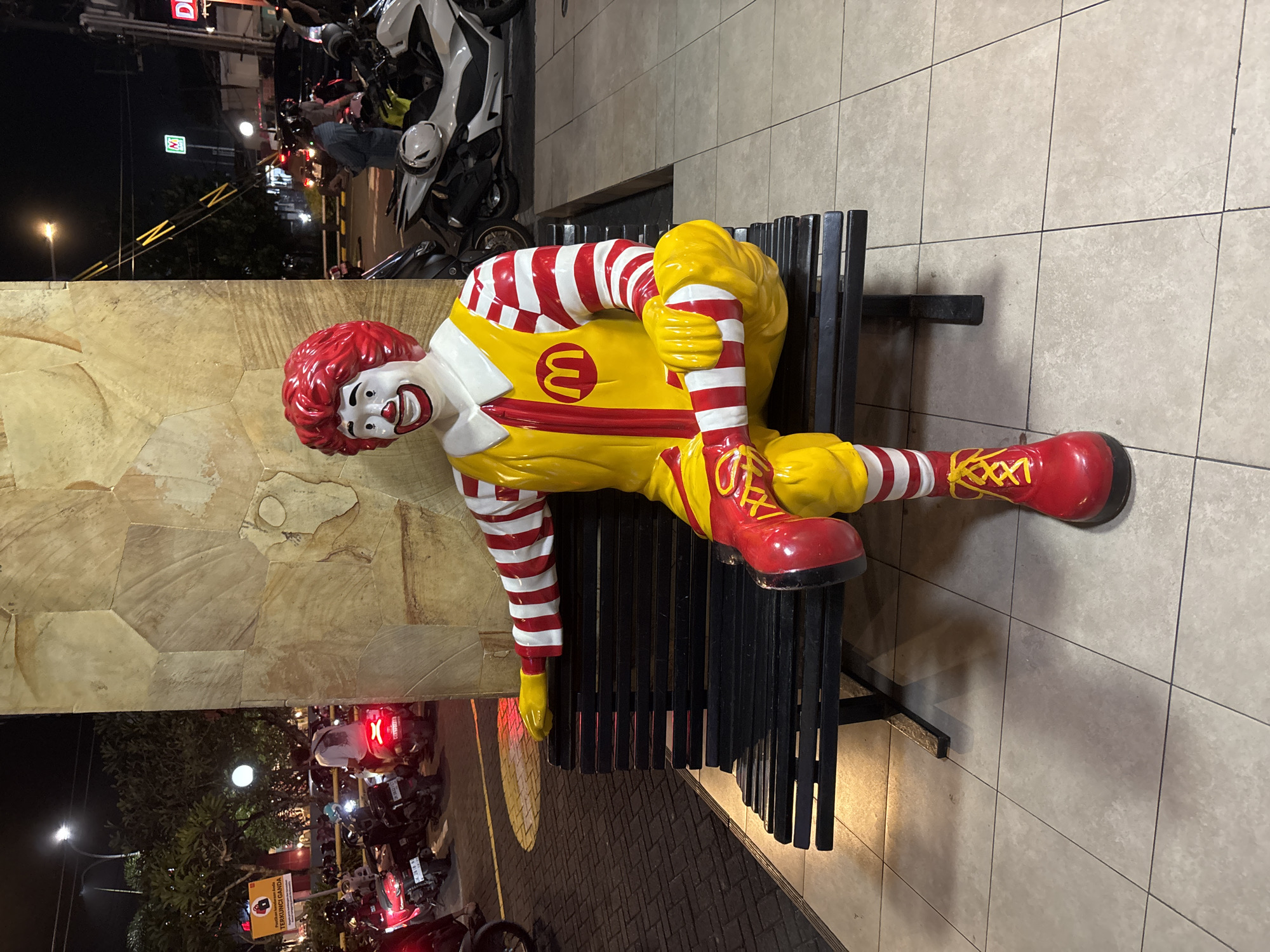 A life-sized statue of Ronald McDonald is prominently featured, sitting on a black slatted bench in an outdoor setting at night. The statue wears a red and white striped shirt, a yellow jumpsuit with a red 'M' logo on the chest, and red boots with yellow laces. Its face is painted with a cheerful, smiling expression, and its red hair is sculpted. The statue is posed with one leg extended and the other bent, arms resting.
The scene is illuminated by artificial lights, indicating nighttime. In the background, behind a patterned stone wall, there's a street or parking area with several motorcycles and scooters, some with riders. Streetlights cast light on trees and a yellow and black striped barrier. The ground around the bench is covered in light-colored square tiles.
Visible text includes the red 'M' logo on Ronald McDonald's costume. In the distance, a green and white sign, identifiable as the 7-Eleven logo, is seen. Further back, a yellow sign with red text partially reads "TERKUNCI GANDA," which is Indonesian for "Double Locked." The location, Kuta Selatan, Indonesia, is characterized by the presence of motorcycles and the local language text.