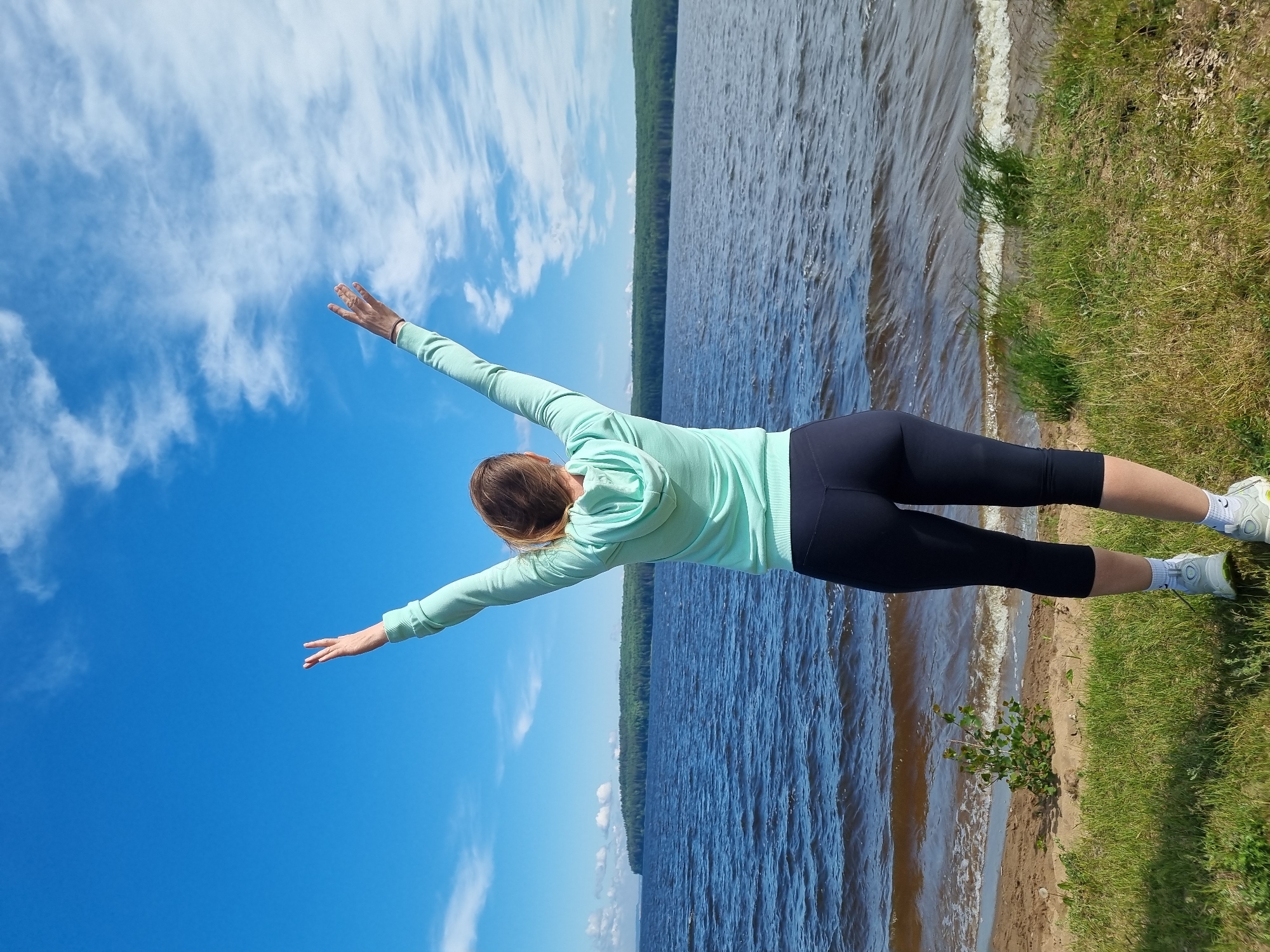 The image, originally oriented sideways, captures a person engaged in a balancing exercise or yoga pose outdoors, seen from behind. The subject, a woman with light brown hair, is wearing a light teal or mint green hooded long-sleeve top and black capri leggings. Her footwear consists of white athletic shoes with white socks. She is balancing on one leg, with her body tilted forward and arms extended horizontally to her sides, resembling a bird in flight or a Warrior III yoga pose. Her head is slightly turned to the right.
The setting appears to be a tranquil natural environment, likely near a large body of water. In what would be the foreground of an upright image, there is a grassy patch giving way to a sandy shoreline. A small green bush grows in the sand near the water's edge. The midground features a wide expanse of deep blue water with ripples on its surface. Beyond the water, in the background, a dense treeline suggests a forest on the distant bank. The sky above is clear and vibrant blue, dotted with scattered white clouds.
The scene conveys a bright, sunny day, likely midday or afternoon, given the clear conditions and strong illumination. There is no visible text in the image. The location context provided is Chaikovsky, Russia.
