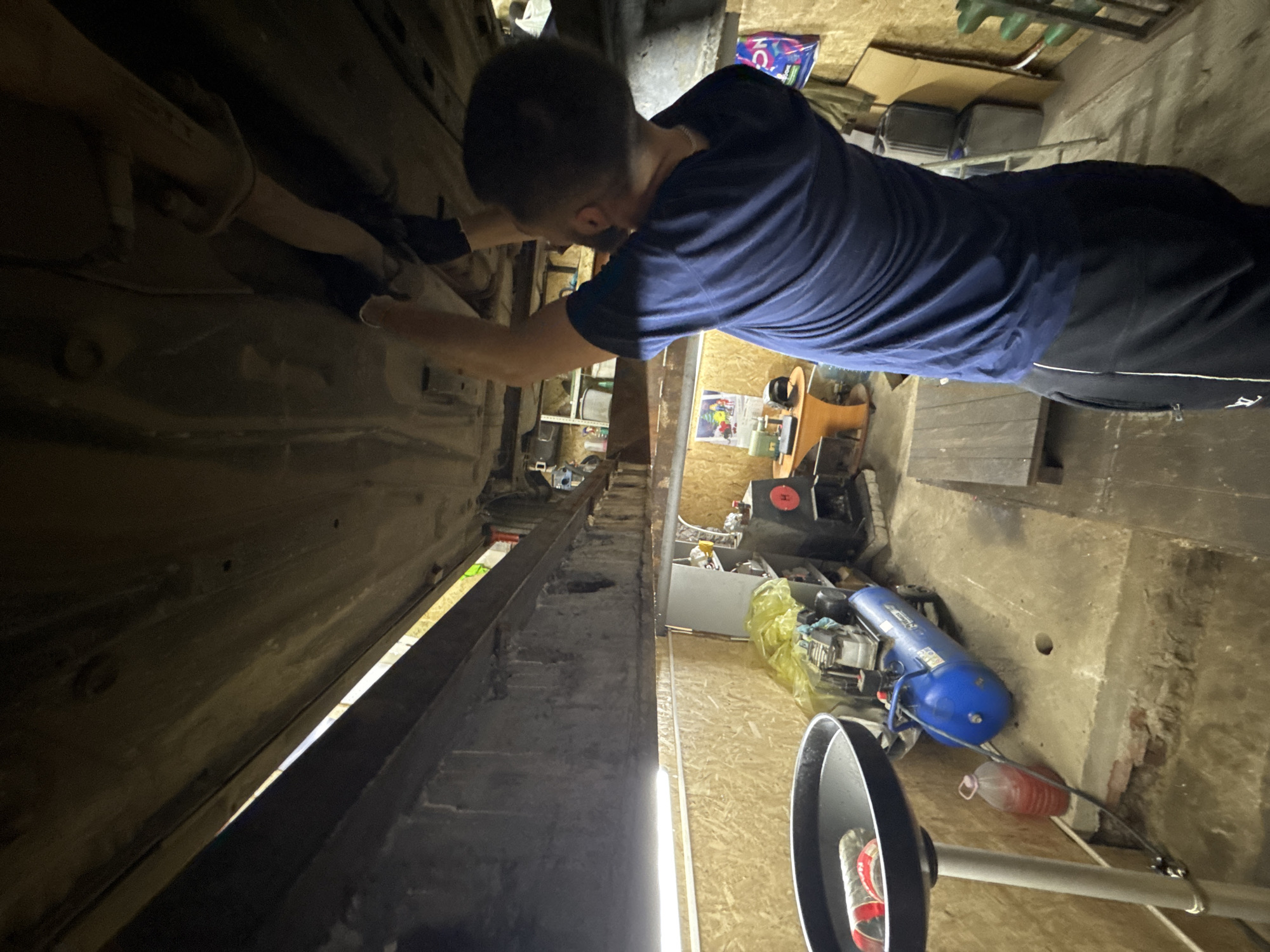 In an automotive workshop located in Perm, Russia, a male mechanic is actively working on the underside of a vehicle elevated on a heavy-duty metal lift. The man, seen from the rear right, wears a dark blue t-shirt and black athletic shorts with a white side stripe and a white logo. His arms are raised, and his gloved hands are engaged with components on the vehicle's dark, grimy undercarriage, which includes exhaust piping and chassis elements. The workshop features light-colored wooden paneling on the walls and a concrete floor, which shows signs of wear and a change in level with exposed brickwork. Overhead fluorescent lighting illuminates the space. Various pieces of equipment are present, including a black oil drain pan on a stand containing a clear bottle to the left, a blue horizontal air compressor on wheels, and shelving units in the background holding tools and miscellaneous items. A small wooden table with a green sewing machine and other objects is visible further back, along with cardboard boxes and a plastic shopping bag on the far right. A wooden pallet rests on the floor to the man's right.