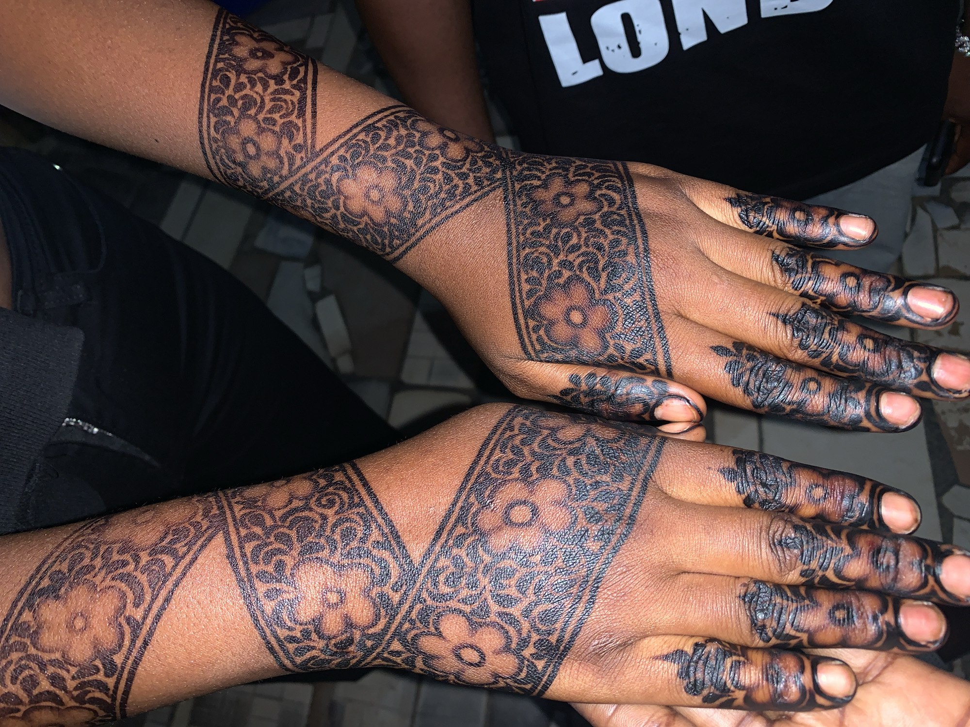 In New Stadium, Nigeria, a close-up shot focuses on the hands and forearms of an individual adorned with intricate henna designs. The dark henna paste is applied in detailed patterns featuring floral motifs and geometric bands. The designs cover the back of both hands, extending up the wrists and forearms, and also wrap around the fingers, reaching the tips. The skin tone is a rich brown. The background is out of focus, showing hints of dark clothing and a tiled surface. The lighting suggests an indoor or shaded outdoor setting.