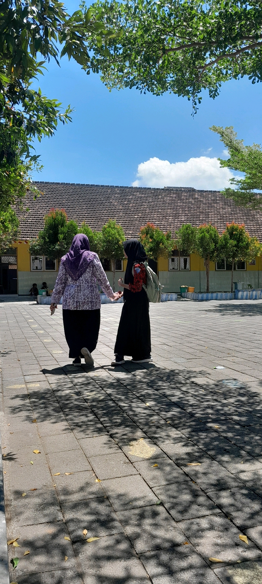 Two individuals, appearing to be female and dressed in hijabs, walk away from the camera, holding hands. The person on the left wears a purple and white patterned top and a black skirt, while the person on the right wears a black abaya and a light-colored backpack. They are walking on a paved courtyard in front of a long, low building with a red-tiled roof and yellow walls. The building has several windows and is flanked by rows of green trees. The sky above is blue with scattered white clouds, and sunlight casts tree shadows on the courtyard. In the background, to the left, two figures are seated. The scene appears to be an outdoor area, possibly a school or community center, consistent with the location of Rogojampi, Indonesia.