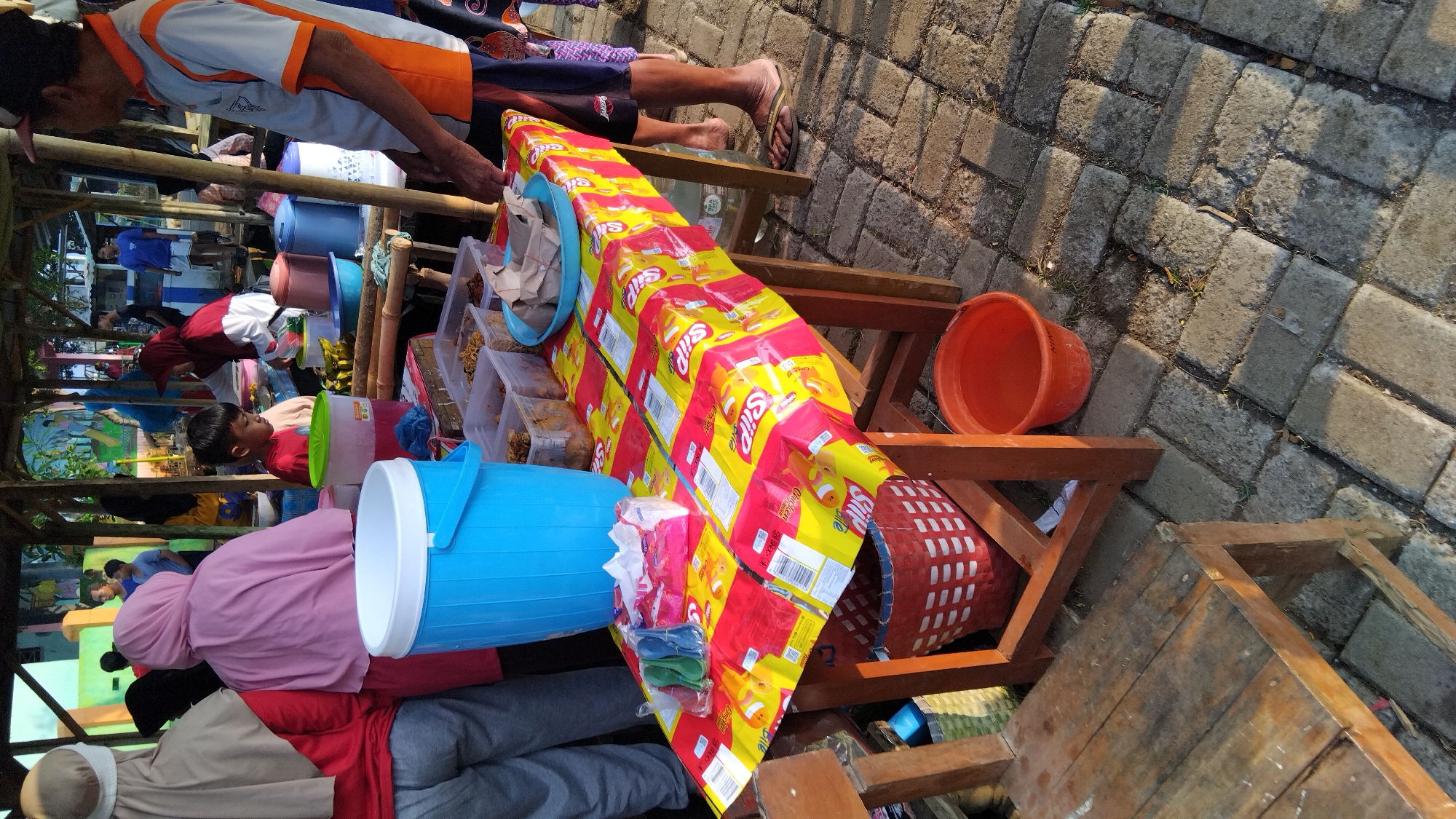 In Sembojo, Indonesia, an outdoor market stall is set up with a wooden table covered by a vibrant red and yellow plastic cloth patterned with images of goldfish and the word "Snack". On the table, there's a large blue cylindrical cooler, plastic containers filled with food, and a blue plastic bowl holding wrapped food items. To the left of the table, a wooden chair is positioned. Behind the table, several people are visible, including a young boy in a red shirt, a woman in a maroon hijab, and other individuals further in the background. The ground is paved with interlocking bricks. To the right of the stall, a person in an orange and white shirt is standing.