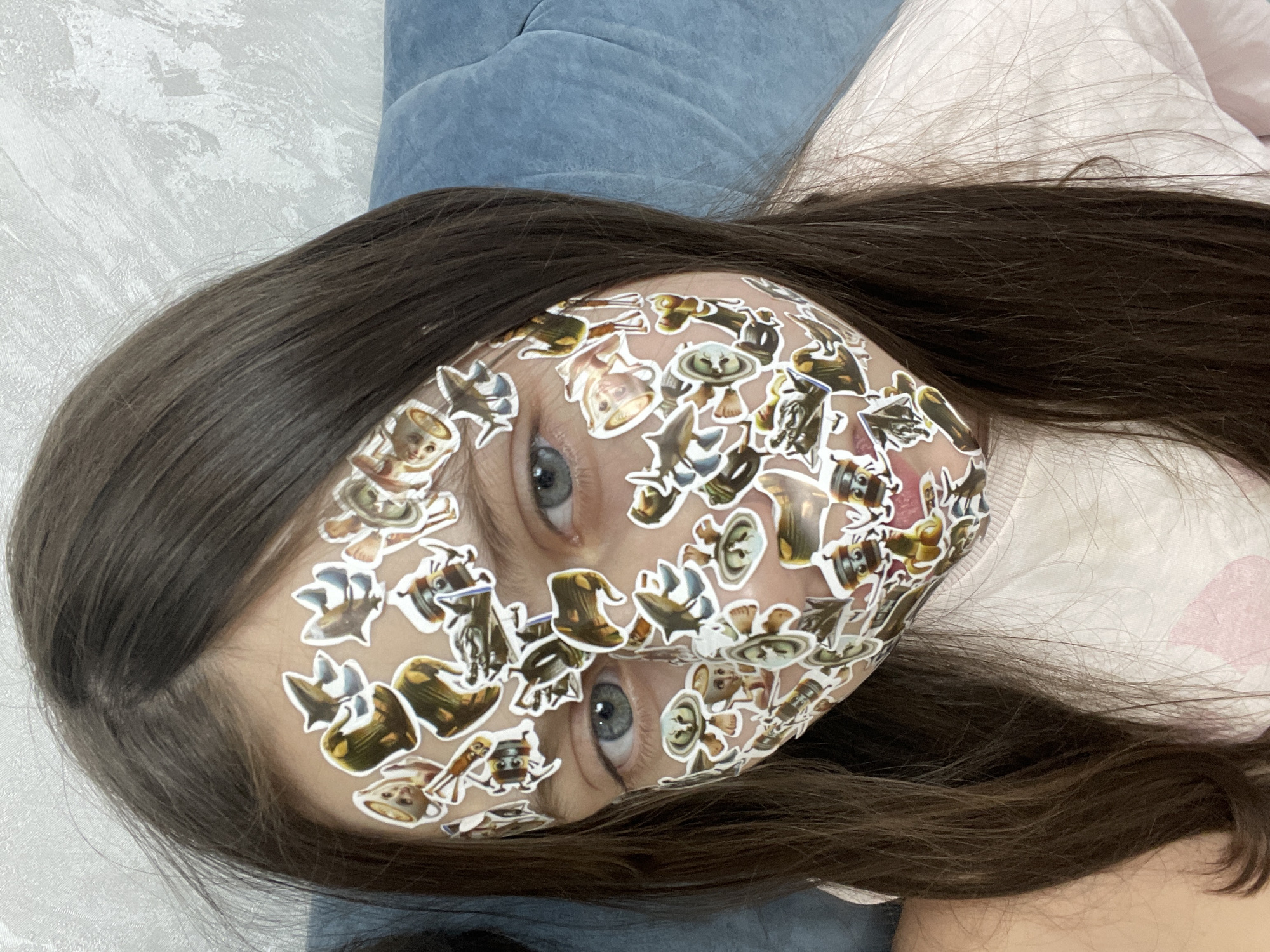 A medium shot depicts a young person lying down, their face covered in stickers. The person's eyes are visible amidst the stickers. The person has long, dark brown hair. They are lying on a surface with a light pink and white patterned fabric visible, with a blue material visible in the upper left corner. No information is provided about the location in Izhevsk, Russia.