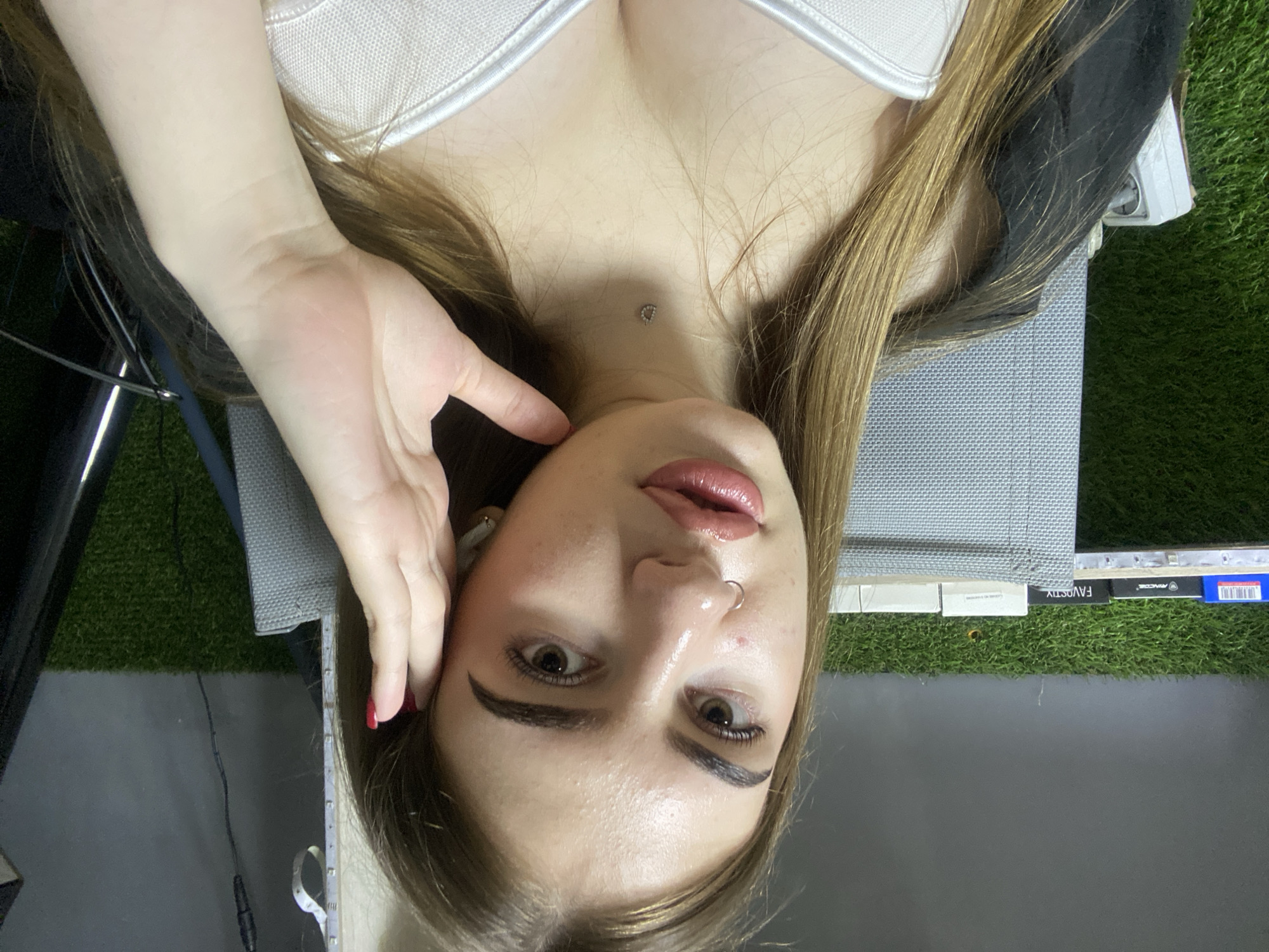A young woman with long brown hair and light skin is looking at the camera. She is wearing a white top and a black jacket. She has a nose ring and a small necklace with a "D" charm. Her left hand is touching her face. In the background, there is a green, artificial grass-like surface and some shelves with boxes. The scene appears to be indoors.