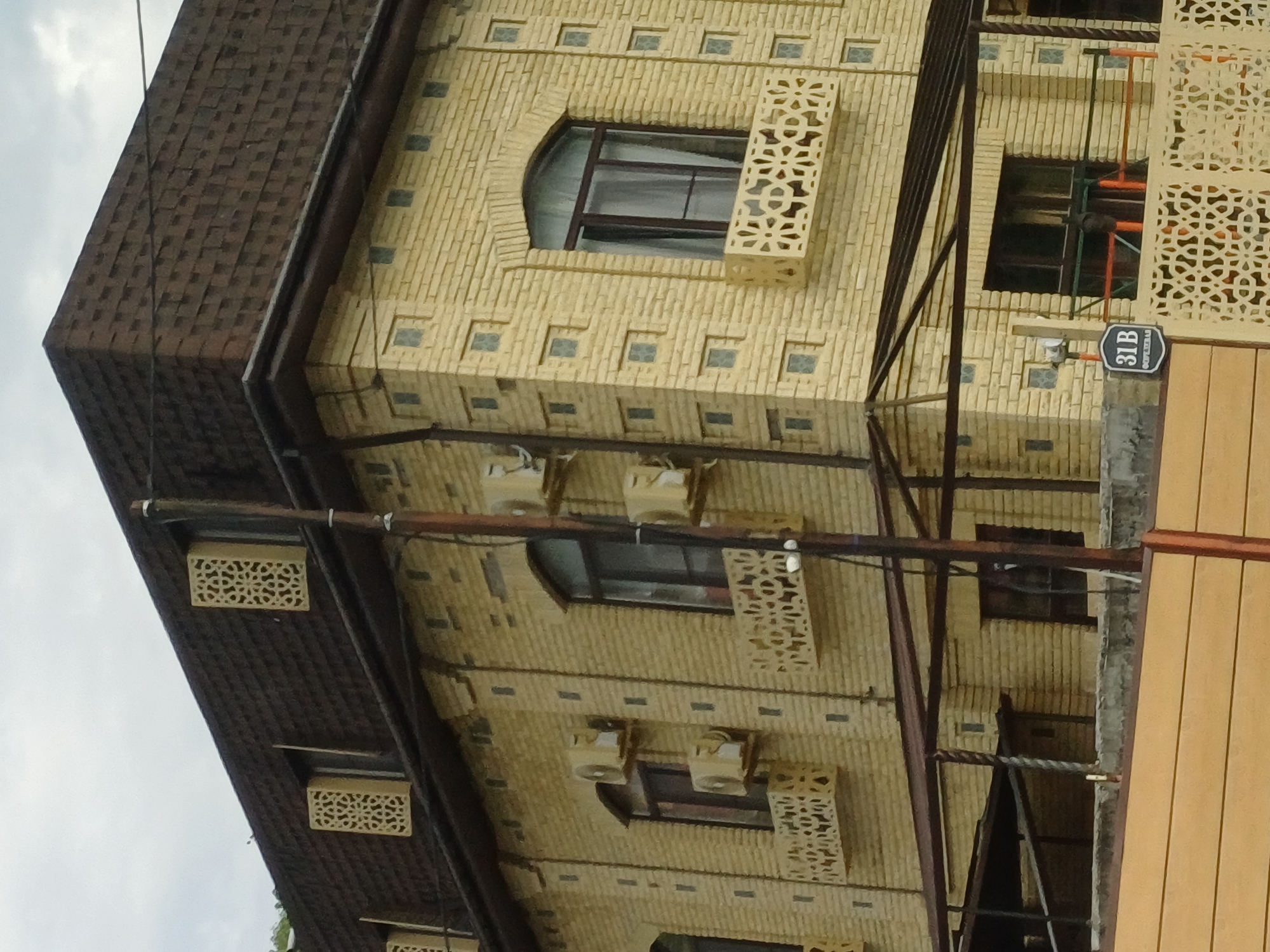 This image captures the exterior of a two-story building constructed with light yellow bricks, located in Kazachiy Brod, Russia. The building features a dark brown asphalt shingle roof with a distinct pyramid-like shape on the upper section. Decorative elements are integrated into the architecture, including ornamental panels above some windows and intricate latticework on small balconies.
The facade is adorned with several air conditioning units mounted on the walls. Windows on the building vary in style, with some being arched and others rectangular. Below the second-story windows, decorative balustrades with a geometric pattern are visible. The building's exterior is also characterized by vertical dark grey rectangular accents and small square windows.
In the foreground, a dark brown support pole stands vertically, with wiring attached to it. Below the second story, a covered outdoor area with a dark brown metal frame and a sloped roof is present. A light wooden fence runs along the bottom of the image. A sign with "31B" and Cyrillic text is affixed to a low wall in front of the building. The sky above is overcast with clouds.