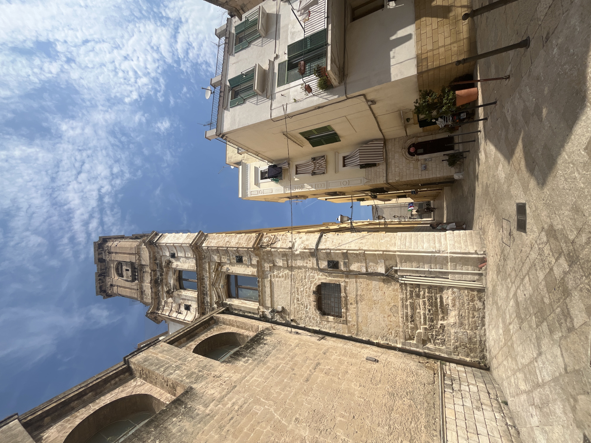 This image captures a narrow, sun-drenched street in the historic district of Bari, Italy, likely in the Old Town (Bari Vecchia). The architecture is predominantly stone, with a tall, ornate bell tower or campanile dominating the left side of the frame, constructed from weathered yellow stone blocks. The tower features arched openings and classical detailing. To its right, a cream-colored building with green shutters and balconies rises, exhibiting a tilted perspective due to the angle of the photograph. The street is paved with large, irregular stone slabs, casting sharp shadows. A few potted plants are visible, including a large orange pot with a green plant near the cream-colored building. In the distance, the alley continues, with more buildings and a glimpse of a few people walking. The sky above is a clear blue with scattered white, wispy clouds. The overall impression is of a charming, ancient European street scene.