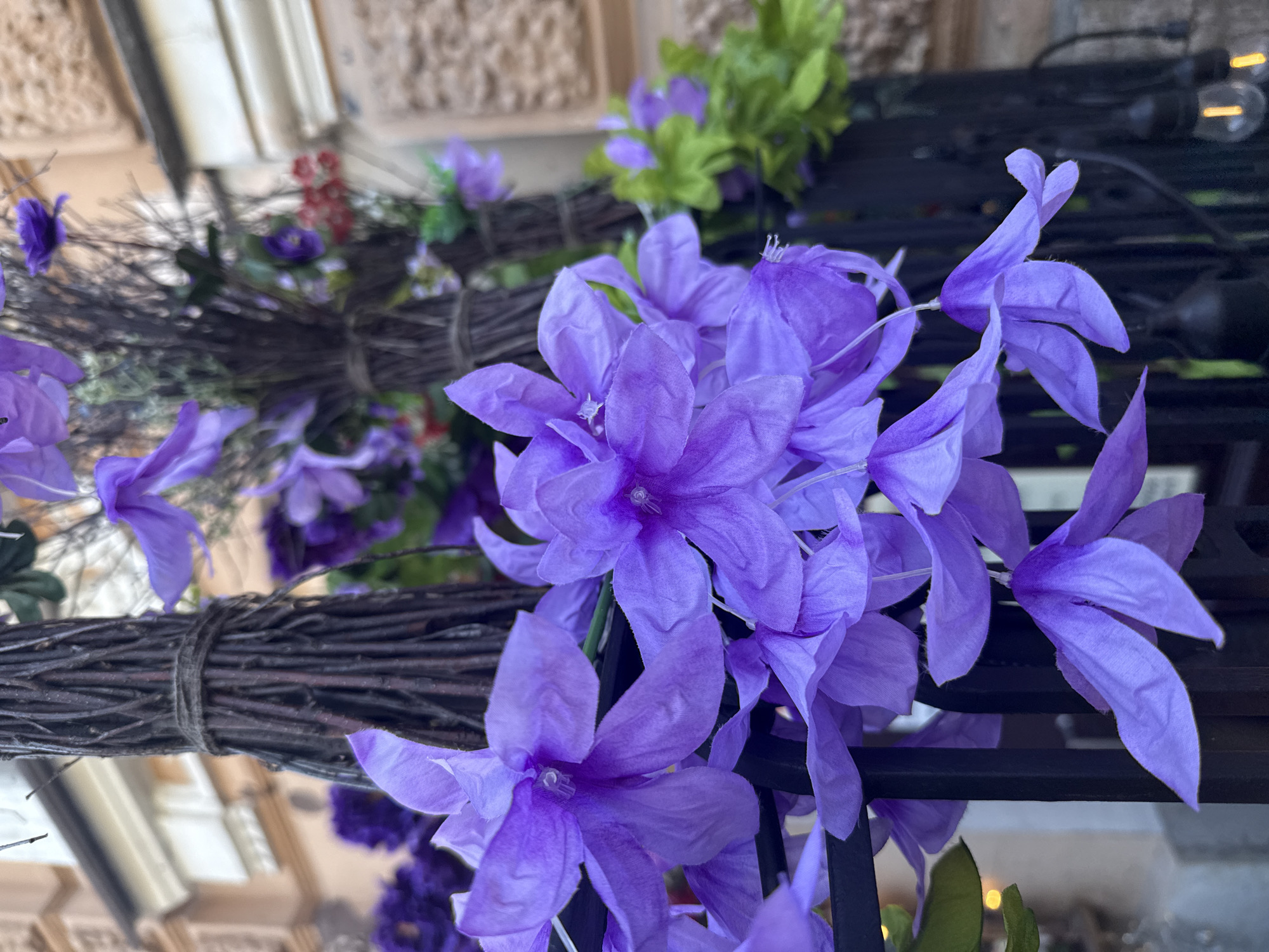 The image shows a close-up view of artificial purple flowers cascading down a black metal fence. In the background, a building with light-colored architectural details is visible, suggesting an urban setting. The overall scene appears to be an outdoor decoration. The specific location in Saint Petersburg, Russia, cannot be determined from the visual information provided in the image.