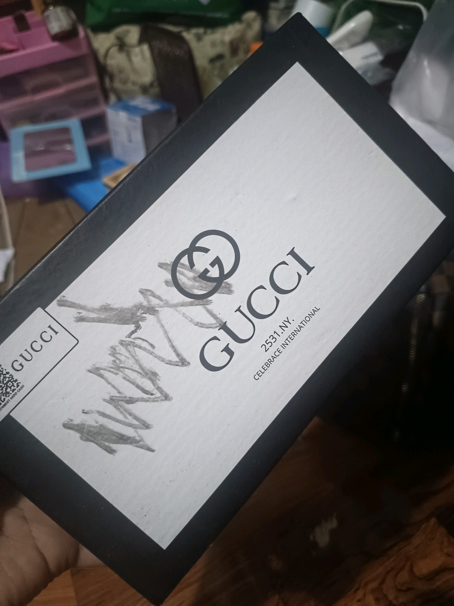 This image shows a close-up of a Gucci box, angled towards the viewer. The box is black with a white lid. The Gucci logo, a stylized double 'G', is prominently displayed in grey. Below it, the word "GUCCI" is printed in black. There is some smudged grey scribbling obscuring parts of the box's surface. To the right of "GUCCI," smaller text reads "2531.NY. CELEBRATE INTERNATIONAL". On the left side of the box, there's a label with "GUCCI" printed vertically, next to a QR code and some smaller text that is mostly unreadable. The box is held by a person's hand, partially visible at the bottom left. The background is out of focus, showing what appears to be a cluttered indoor environment with shelves and various objects. There is no visible indication of the location being Masintan, Indonesia.