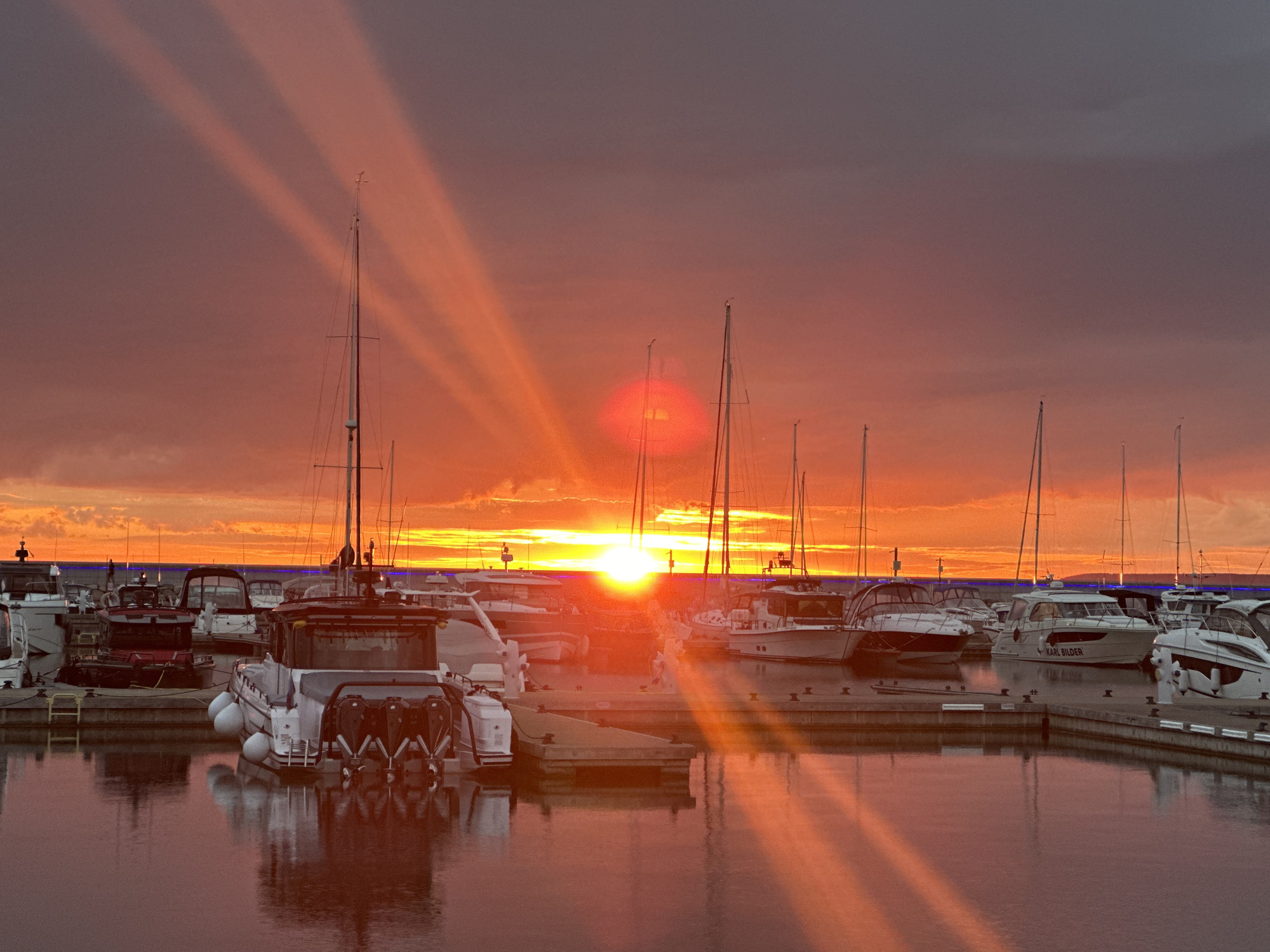 This photograph captures a vibrant sunset over a marina in Tallinn, Estonia. The sky is a dramatic canvas of fiery oranges, pinks, and deep purples, with distinct rays of sunlight piercing through the clouds, creating a striking visual effect.
The marina is filled with numerous docked boats, predominantly sailboats and some motorboats. The masts of the sailboats create a linear pattern against the colorful sky. Several boats are clearly visible, including a white motorboat with "KARL BILDER" written on its side. The reflections of the boats and the sunset are beautifully mirrored in the calm water of the marina.
The scene suggests a peaceful evening at the end of the day. The sun is low on the horizon, indicating either sunrise or sunset, but the intensity of the colors and the long rays point towards sunset. The weather appears clear, with the dramatic cloud formations adding to the picturesque quality of the sky. The overall mood conveyed is one of tranquility and natural beauty, enhanced by the serene setting of the marina.