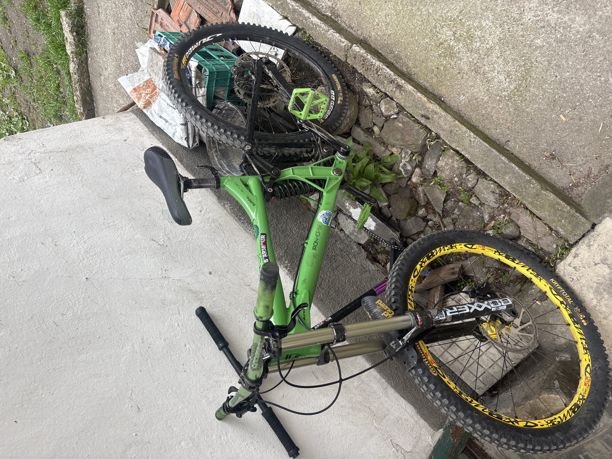 A green mountain bike lies on its side next to a concrete surface in Devin, Bulgaria. The bike is lying on top of some rocks, dirt, and a few weeds. Its wheels are black and yellow. The handlebars are black.