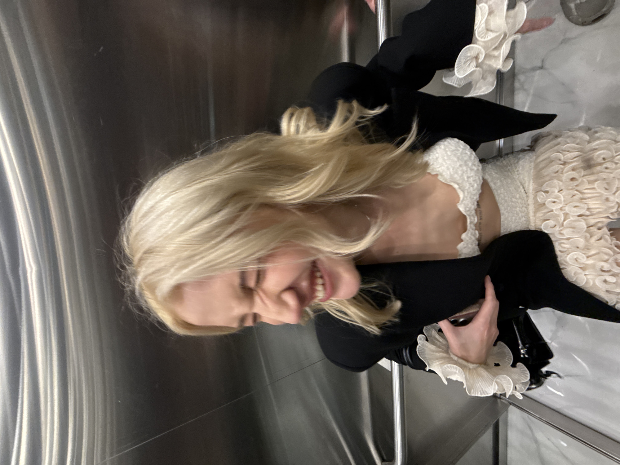 A woman with blonde hair is smiling and appears to be inside an elevator. She is wearing a white dress with ruffled details and a black blazer with similar ruffled embellishments on the cuffs. The elevator has brushed metal walls. The location and time of day are not discernible from the image. The city of Belgrade, Serbia, cannot be confirmed as the location.