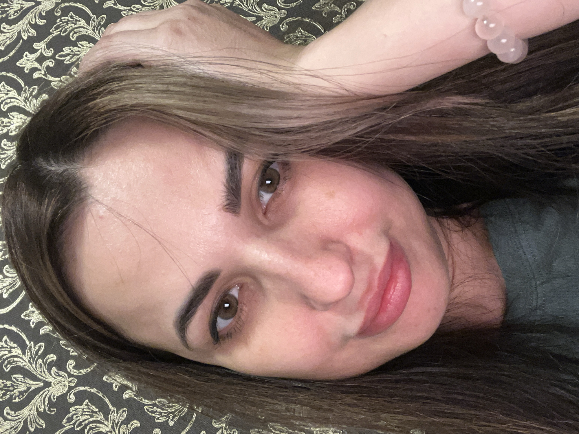 A close-up portrait of a woman with long brown hair and brown eyes. She is lying on a patterned surface, possibly a bed or decorative fabric. Her left arm is raised and bent, with a beaded bracelet visible on her wrist. The location is Unknown, Unknown.