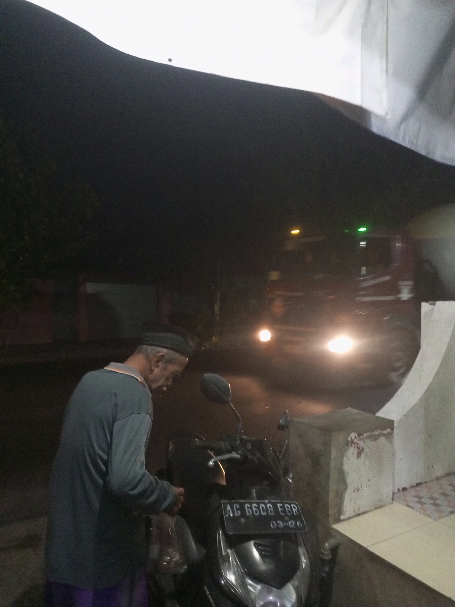A dimly lit scene, possibly in the city of Ngasem, Indonesia, captures an elderly man in a dark green long-sleeved shirt and a dark cap standing next to a black motorcycle. He is holding a clear plastic bag with items inside. The motorcycle's license plate reads "AG 6608 EBR" with a date stamp below. In the background, a large truck with its headlights on is partially visible, suggesting it is either parked or moving slowly. The environment appears to be outdoors, with some foliage and a building facade on the right. The overall lighting suggests it is nighttime.