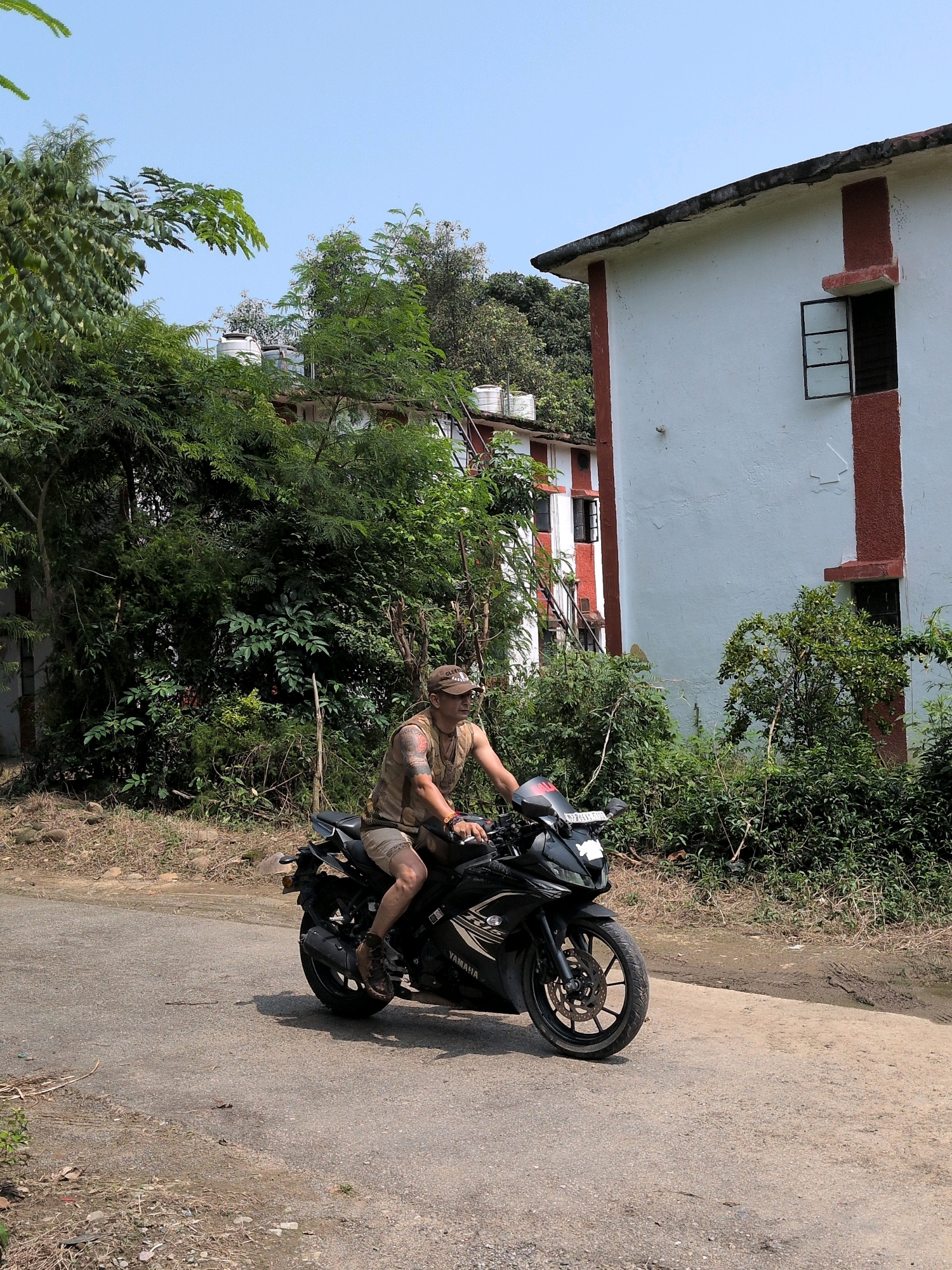 A person is riding a black Yamaha motorcycle on a paved road. The rider is wearing a camouflage vest, shorts, a baseball cap, and brown shoes. They have tattoos on their left arm and are looking to the right. The motorcycle has a license plate. The road is surrounded by lush green trees and vegetation. In the background, there are buildings with white and red accents, and a clear blue sky. The scene appears to be outdoors, possibly in a rural or semi-urban area of Pathankot, India, given the vegetation and building style.