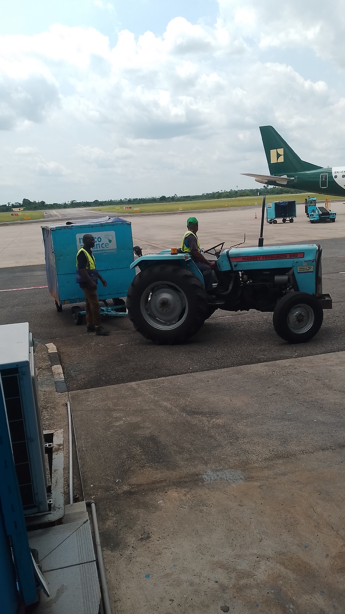 The image depicts an active ground scene at an airport, likely in Orerokpe, Nigeria, under a partly cloudy sky during daylight hours. In the foreground and midground, two airport ground staff are engaged in moving cargo.
One man, wearing a lime green high-visibility vest over a dark shirt and dark trousers, is driving a light blue Massey Ferguson 240 tractor. He is positioned on the left side of the tractor's seat, looking forward. The tractor has large rear wheels and smaller front wheels, and part of a "CARGO SERVICE" decal is visible on its side. The second man, also in a lime green high-visibility vest over a dark shirt and brown trousers, stands beside a light blue cargo container. He appears to be guiding or securing the container, which is attached to the tractor via a tow bar. Text visible on the cargo container reads "eco Finance" and partially "www.mahrofinance.com".
In the background, the tail section of an airplane is visible. The tail is dark green with a yellow, possibly square-shaped, logo near the top. Part of the aircraft's registration, "5N-EN", is discernable on the fuselage. Further in the background, another blue ground support vehicle with multiple wheels and a smaller blue unit are parked. Beyond the tarmac, a runway with visible markings like "30-12A" and "20-04A" stretches into the distance, flanked by grassy areas and distant greenery under the expanse of clouds. The ground in the immediate foreground consists of textured concrete with some pipes and an air conditioning unit structure.