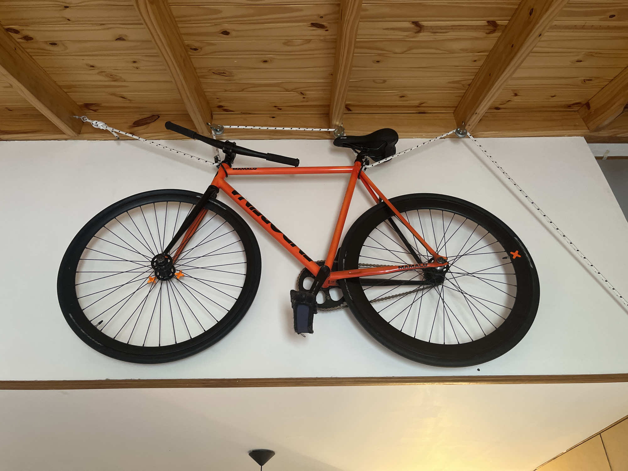 An orange and black fixed-gear bicycle hangs suspended from the ceiling and a wall. The bicycle is suspended by a white rope system attached to the ceiling via pulleys. The rope is also attached to the handlebars and seat of the bike. The bicycle is hanging against a white wall, above a light brown wainscot. The ceiling is made of wood planks. The location is unknown, and cannot be determined to be Ushuaia, Argentina.