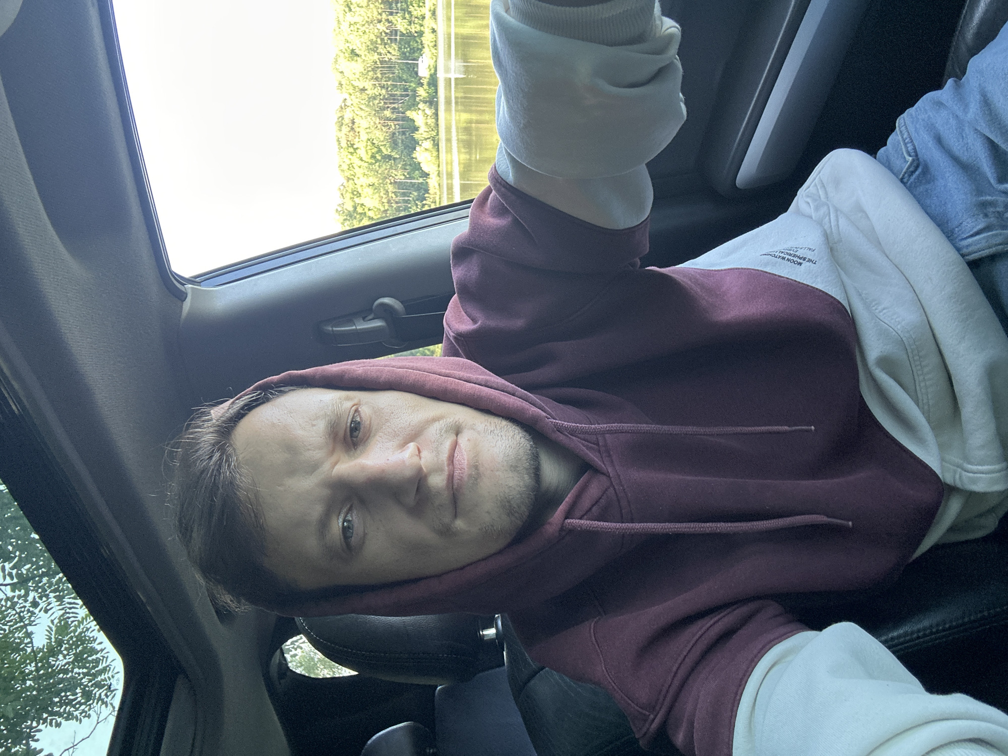 A man is sitting in the driver's seat of a car, looking at the camera. He is wearing a maroon hoodie with cream-colored sleeves and blue jeans. The car's sunroof is open, and trees and a body of water are visible in the background. The scene appears to be in a natural setting, possibly near Makiv, Ukraine, given the greenery and water. The man's expression is neutral.