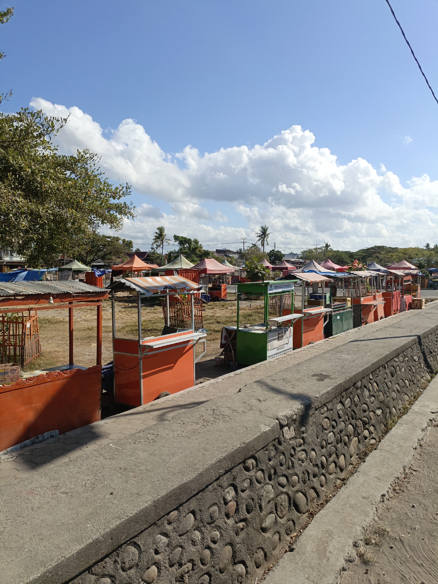 The image depicts an outdoor market scene in Bulukumba, Indonesia, likely located near a coastal area, suggested by the presence of a stone-lined drainage ditch that could be a canal or part of a waterfront development. The market consists of numerous small, colorful stalls, many of which are empty. The stalls have orange, green, and red awnings and fronts, with some displaying simple tables and structures. The ground is covered in dry grass and dirt. In the background, trees, buildings, and palm trees are visible, indicating a tropical or subtropical climate. The sky is a vibrant blue with fluffy white clouds, suggesting a sunny day. The foreground features a concrete walkway or embankment along the side of the market, with a stone-lined ditch to its right. No people are visible in the market stalls.