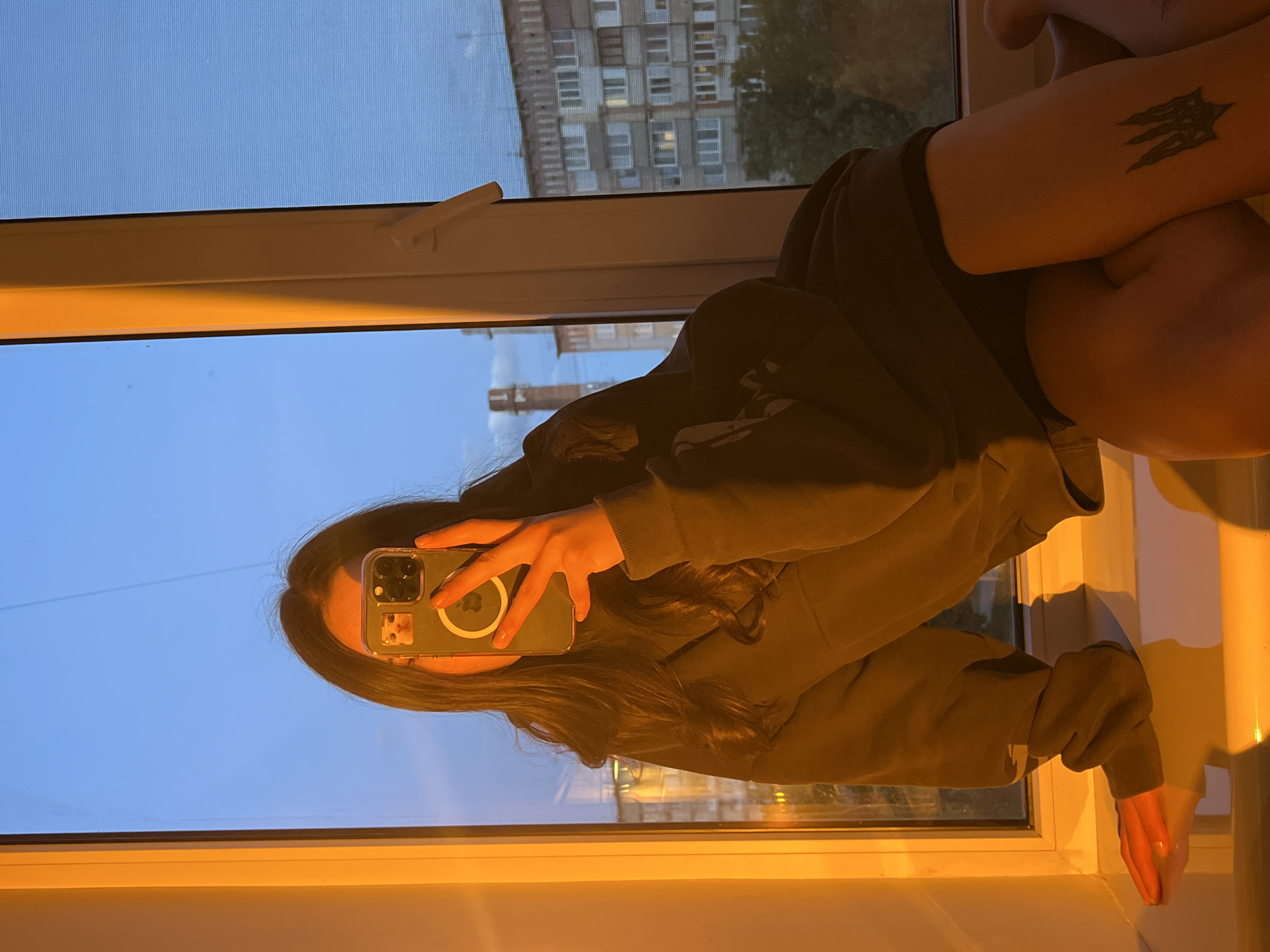 A woman is sitting on a windowsill, taking a selfie with her phone. The phone is held up in front of her face. The woman is wearing a hoodie. She has long, dark hair. Her elbow, with a tattoo, rests on her knees. Outside the window, a multi-story building can be seen. Based on the tattoo on her arm and building design, the location is most likely in Ternopil, Ukraine. The time of day is dusk or dawn.