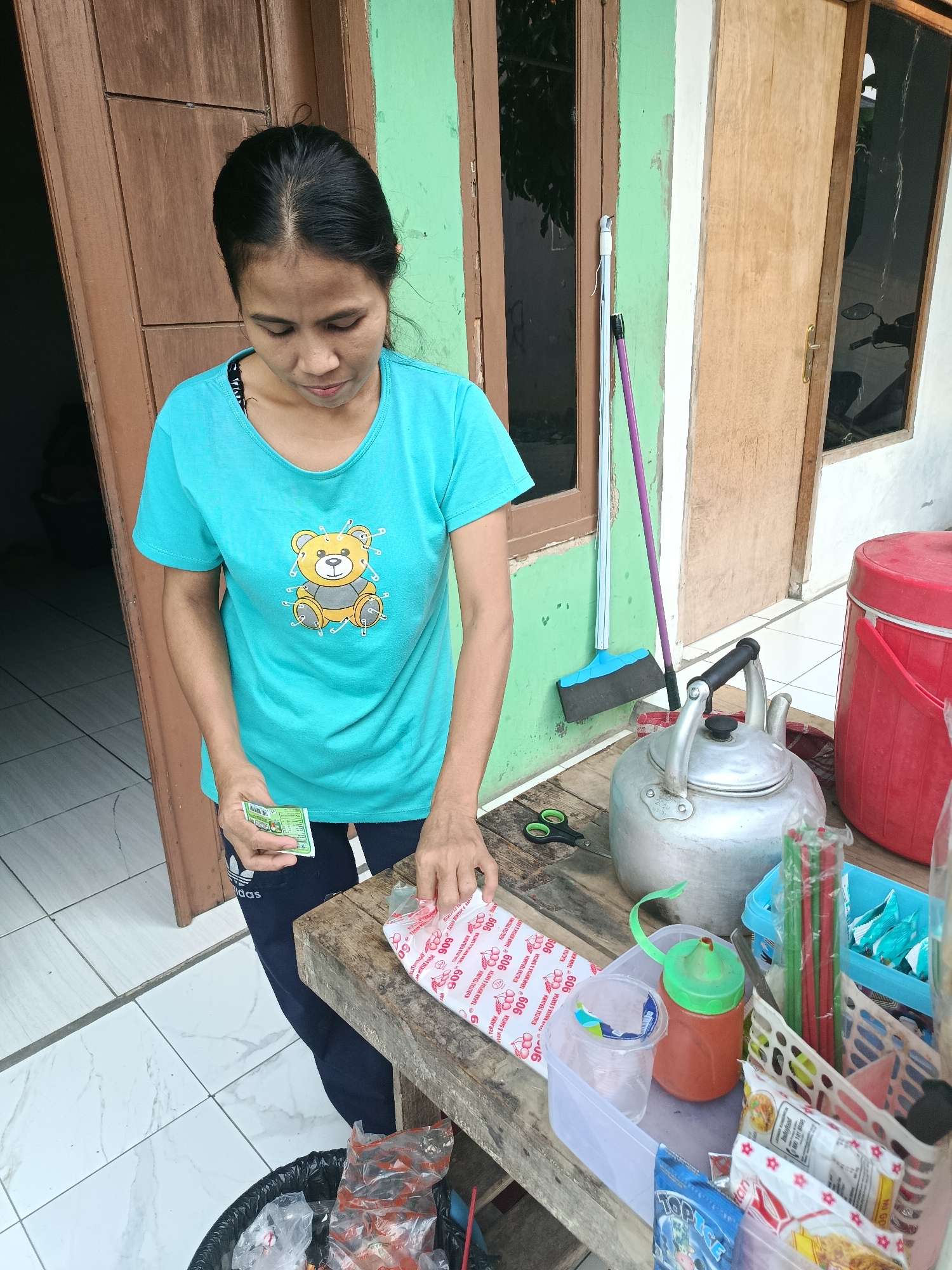 An Indonesian woman, with black hair pulled back and wearing a turquoise t-shirt with a teddy bear graphic, stands in front of a weathered wooden and green painted building. She is holding a small green packet in her left hand and is in the process of opening a long, white, individually wrapped packet of food items with red lettering in her right hand. She is wearing navy blue athletic pants with white stripes.
To her right, on a rustic wooden table, are various items: a silver metal kettle with a black handle, a green plastic condiment bottle with a red dispenser top, a clear plastic cup, and a blue basket filled with red and green straws. Behind these items, a large red cylindrical container with a white lid is visible.
In the background, a broom with a blue and black head and a purple handle leans against the green wall. The floor is tiled with a white and grey pattern. The overall scene appears to be an outdoor area, possibly a small shop or food stall, on a street in Balaraja, Indonesia.
