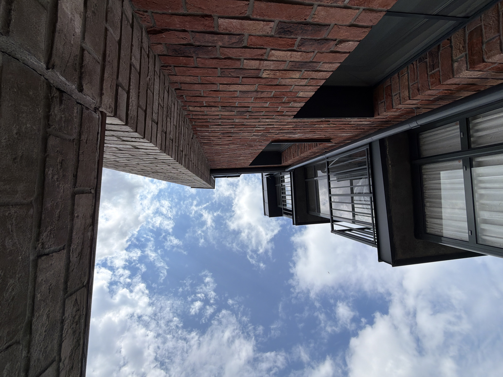 The image depicts an upward view of a building facade with a brick exterior and a balcony against a blue sky with white clouds. The building appears to be modern, with dark-framed windows and a metal railing on the balcony. The brickwork is a prominent feature, showcasing shades of red and brown. One portion of the building's upper edge is visible, with a different type of brick or stone material. No people are visible in the scene. The location in Strelna, Russia, is not discernible from the visual elements alone; it is an inferred piece of information.