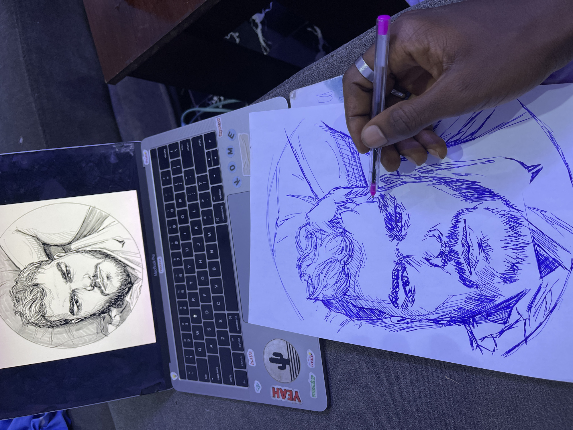 This media file captures a close-up view of a person's hand holding a pink pen and drawing a portrait on a sheet of paper. The paper is placed on top of a MacBook Pro laptop, suggesting the drawing is being done from a reference image displayed on the screen, which shows a sketch of a man's face within a circle. The drawing on the paper is in progress, with bold blue lines sketching out a portrait. The laptop's keyboard is partially visible, and it has several stickers on its palm rest, including one with a cactus and text that reads "hustle," "YEAH," and "create."
The scene appears to be indoors, with the lighting suggesting it might be evening or a dimly lit room. The subjects are a person's hand, the laptop, and the ongoing drawing. The activity is the creation of art, specifically a portrait sketch.
Notable details include the silver ring on the person's ring finger. The stickers on the laptop provide some context about the person's interests or aspirations, hinting at a creative or entrepreneurial mindset. While no specific time of day or weather can be determined, the overall mood feels focused and artistic. The context provided states the location is Abuja, Nigeria. No discernible emotions are explicitly shown, but the act of drawing often implies concentration and creativity.