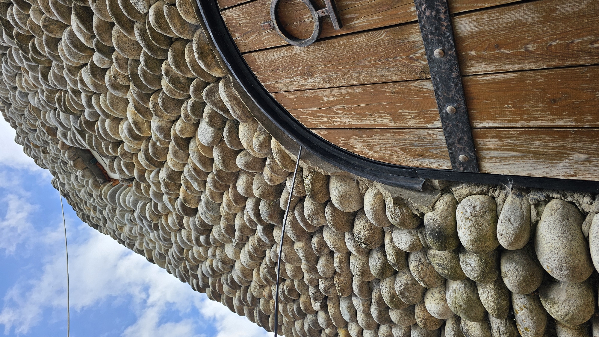 A low-angle, medium shot captures the textured wall of a round building constructed from tightly packed, smooth, gray and tan river stones. The wall extends upwards and to the left, meeting a cloudy, pale blue sky. A thin, off-white wire stretches horizontally across the upper left portion of the frame, disappearing behind the stone wall. Partially visible on the right side of the image is a large, arched wooden door with weathered, vertical planks and dark metal banding. The door features a decorative circular metal emblem with a horizontal bar. The scene appears to be an exterior architectural detail, possibly in Красный Курган, Russia, given the contextual information.