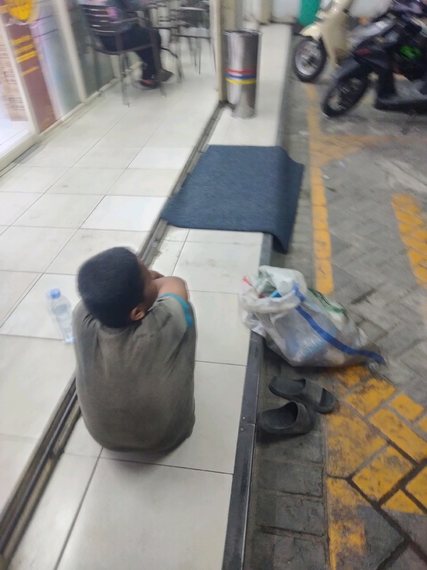 In Pisangan Jaya, Indonesia, a child is seen from behind, sitting on a tiled floor with their back to the camera and head tucked into their arms. The child is wearing a grey shirt with blue trim and is nestled within a large grey fabric sack. To the child's left, a clear plastic water bottle with a blue cap rests on the floor. A dark blue mat is laid out on the tiles in front of the child, and a metal trash can with colored bands is visible further back. Beyond the mat, the ground is paved with concrete and marked with yellow lines, with two black sandals and a plastic bag filled with items lying on it. In the background, a person is seated at a table with chairs, and two motorcycles are parked. The environment appears to be an outdoor area adjacent to a building with glass windows.