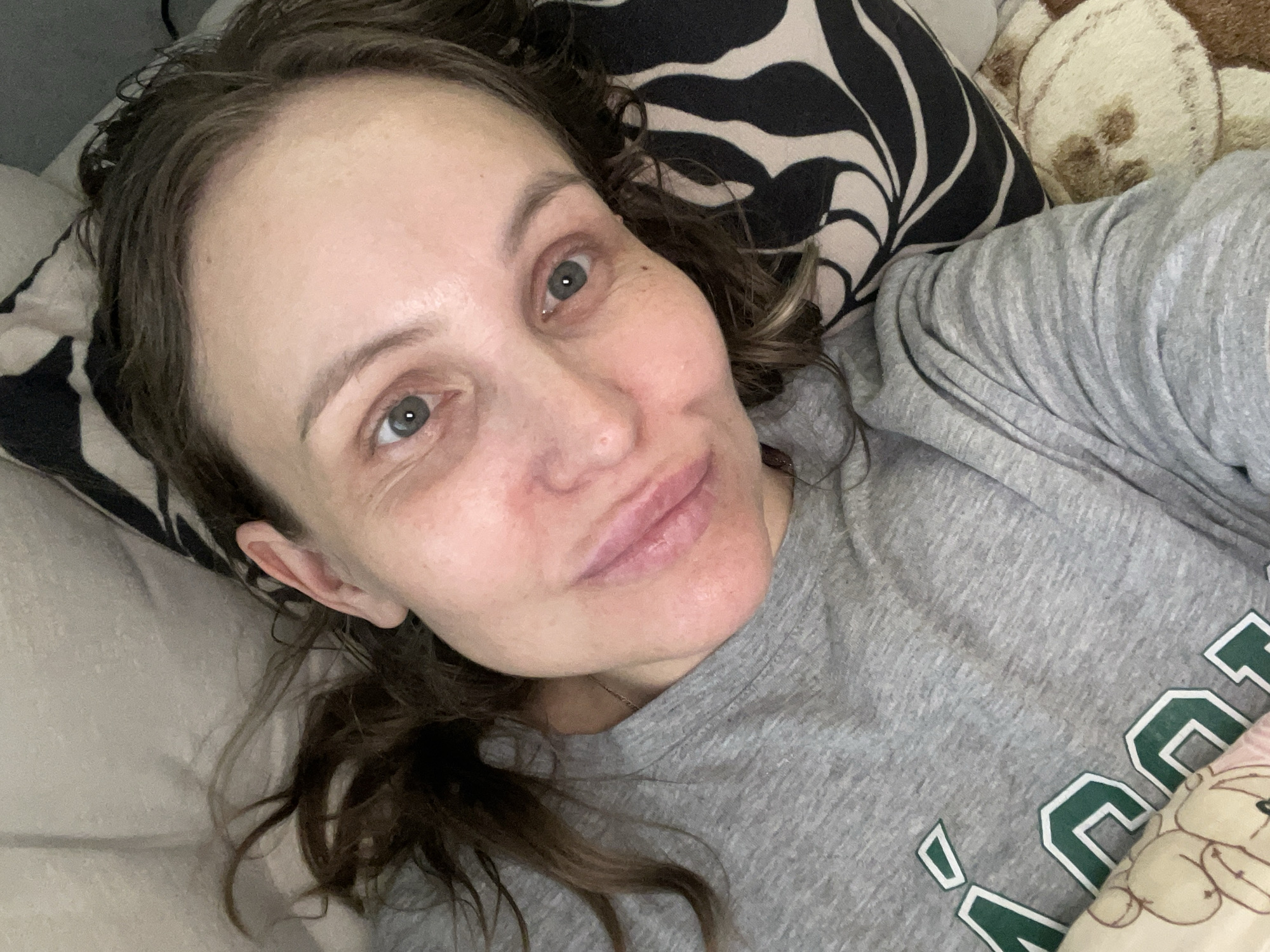 A woman is lying down, taking a selfie. She is wearing a grey shirt with green and white lettering on it that says "AGU." She is lying on a bed with a white pillow to the left and pillows with a zebra-like pattern and a teddy bear pattern behind her. Her hair is brown and slightly disheveled. Her eyes are light-colored. The location is not discernible from the image.