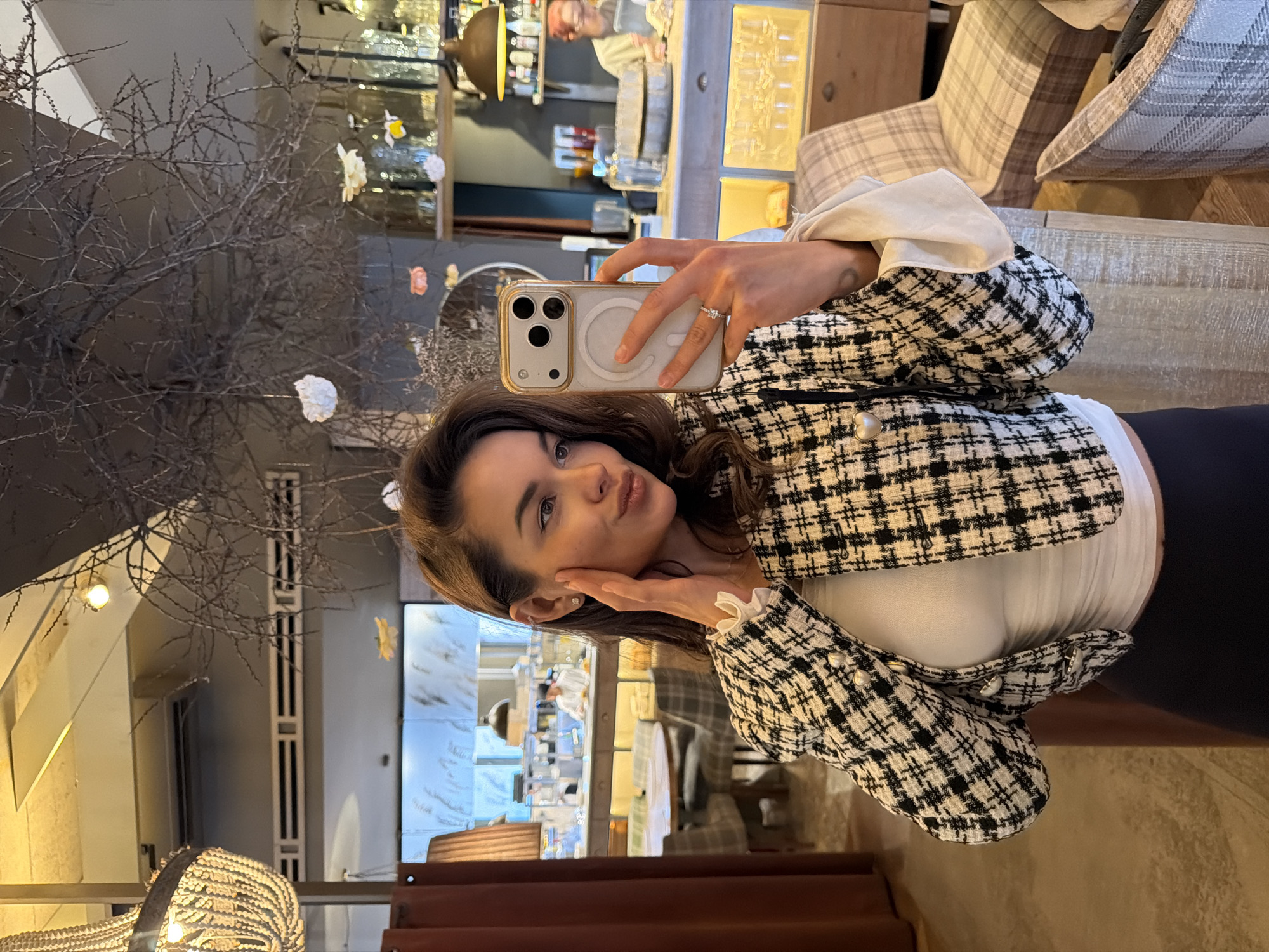 A woman is taking a selfie indoors. She is holding a white smartphone up to her face with her left hand and pouting her lips. She is wearing a black and white checkered jacket with white ruffles on the sleeves, a white top, and black pants. She is wearing a ring on her left ring finger and a small earring in her left ear. The background includes a bar area with various bottles and glasses on shelves, as well as a plaid armchair. A bare tree branch with small white paper flowers is visible on the left. The location may be in the city of Krasnoyarsk, Russia.