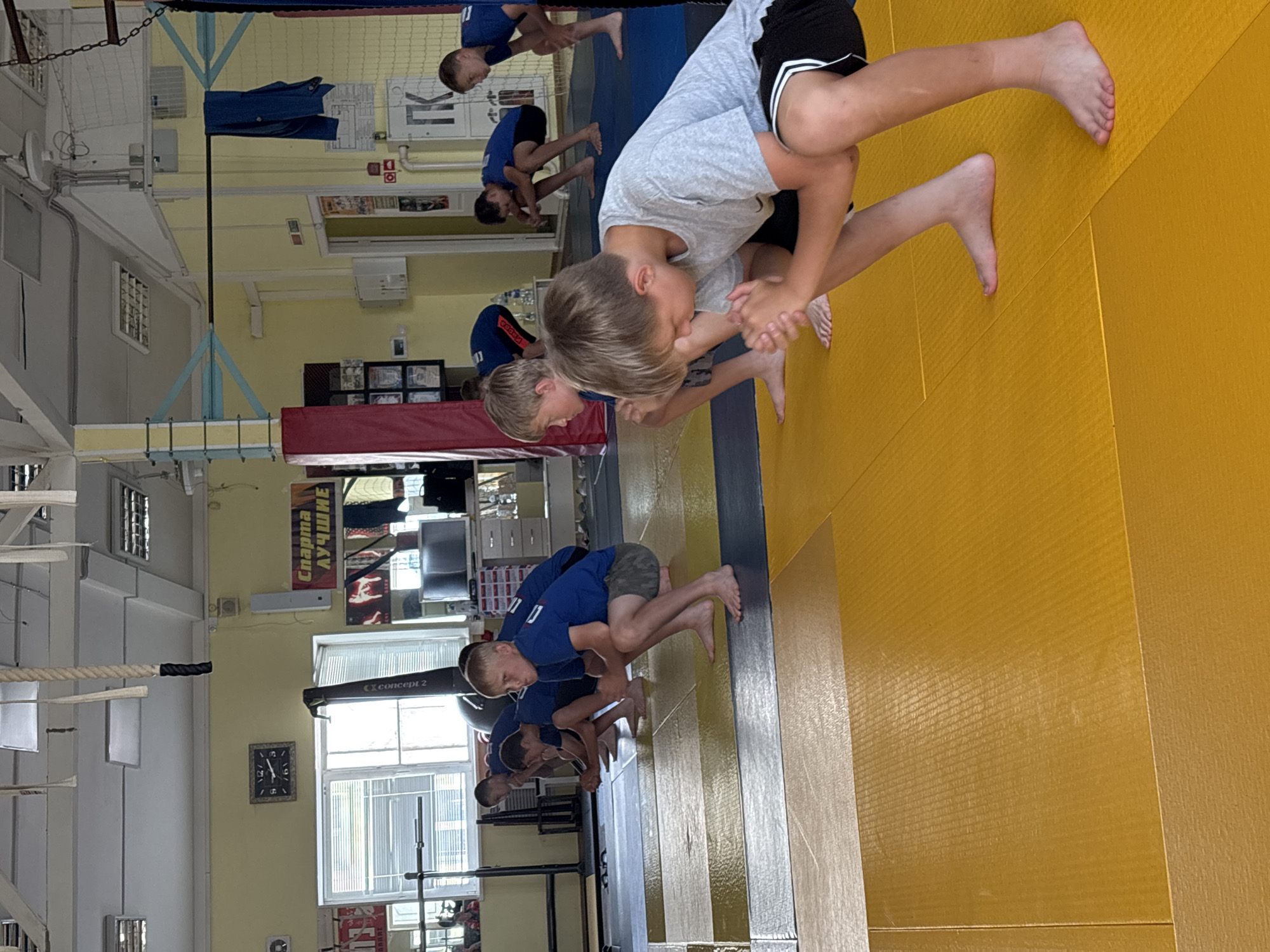 The image shows a group of young boys practicing judo on a yellow mat in what appears to be an indoor training facility. The boys are wearing judogi, which are traditional Japanese martial arts uniforms, or t-shirts and shorts. They are in various grappling positions, demonstrating physical activity and training.
In the background, there are elements of a gym or sports hall, including climbing ropes, a clock on the wall showing a time close to 12:20, and windows. There are also posters on the wall, one of which has Russian text that appears to read "Спорт - лучший" which translates to "Sport is the best". Another poster has a logo that resembles "concept 2".
The scene is well-lit, suggesting it is daytime. The overall atmosphere seems to be one of focused training and physical exertion. The image is oriented sideways, making it appear as if the subjects are on a wall.