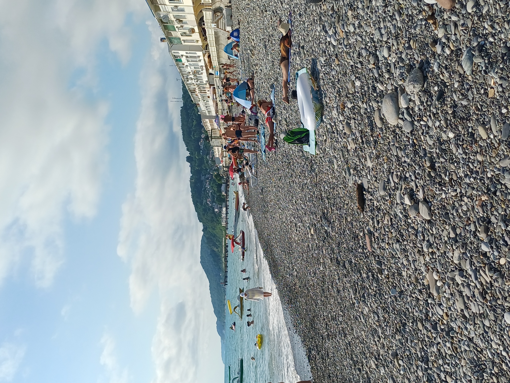 This is a wide-angle, eye-level, vertical shot of a crowded pebble beach in Sochi, Russia, on a sunny day. The scene is dominated by a long, multi-story building with many windows and balconies lining the edge of the beach. Below the building, a dense collection of people are sunbathing on towels and mats, many wearing swimsuits. Some are seated, while others are lying down. Umbrellas are interspersed among the sunbathers, providing shade.
The beach itself is covered in smooth, gray pebbles. The sea is visible to the left, with gentle waves lapping the shore. Several people are in the water, some swimming and others wading. Inflatable toys, including a ring and a yellow float, are scattered in the water. A red kayak is also visible. The background shows a green, tree-covered mountainside rising behind the building, with a cloudy sky above.
The time of day appears to be afternoon, indicated by the bright sunlight and the shadows cast by the building. The weather is clear and warm, suitable for a day at the beach. The overall mood is one of relaxation and recreation, with many people enjoying the summer weather. There is no visible text in the image.