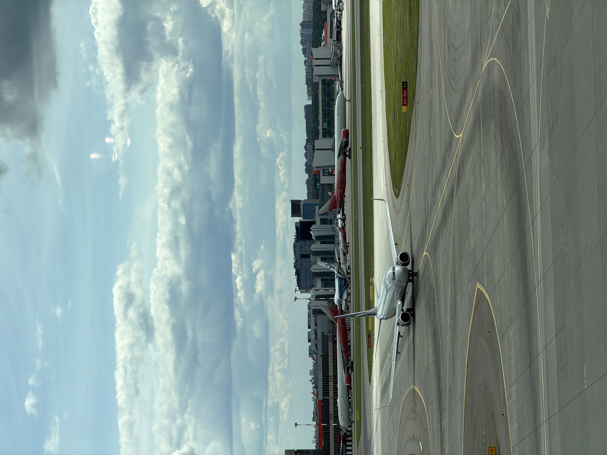 The image shows an airport scene with multiple airplanes on the tarmac. A silver private jet is in the foreground, taxiing on the runway. Other larger commercial airplanes, some with red and white livery, are parked at gates connected to terminal buildings in the middle ground. In the background, a cityscape with high-rise buildings and a cloudy sky is visible. The scene is set at Sheremetyevo International Airport located near the city of Khimki, Russia, indicated by the architectural style of the terminal buildings and the surrounding landscape. There are runway markings and signage.