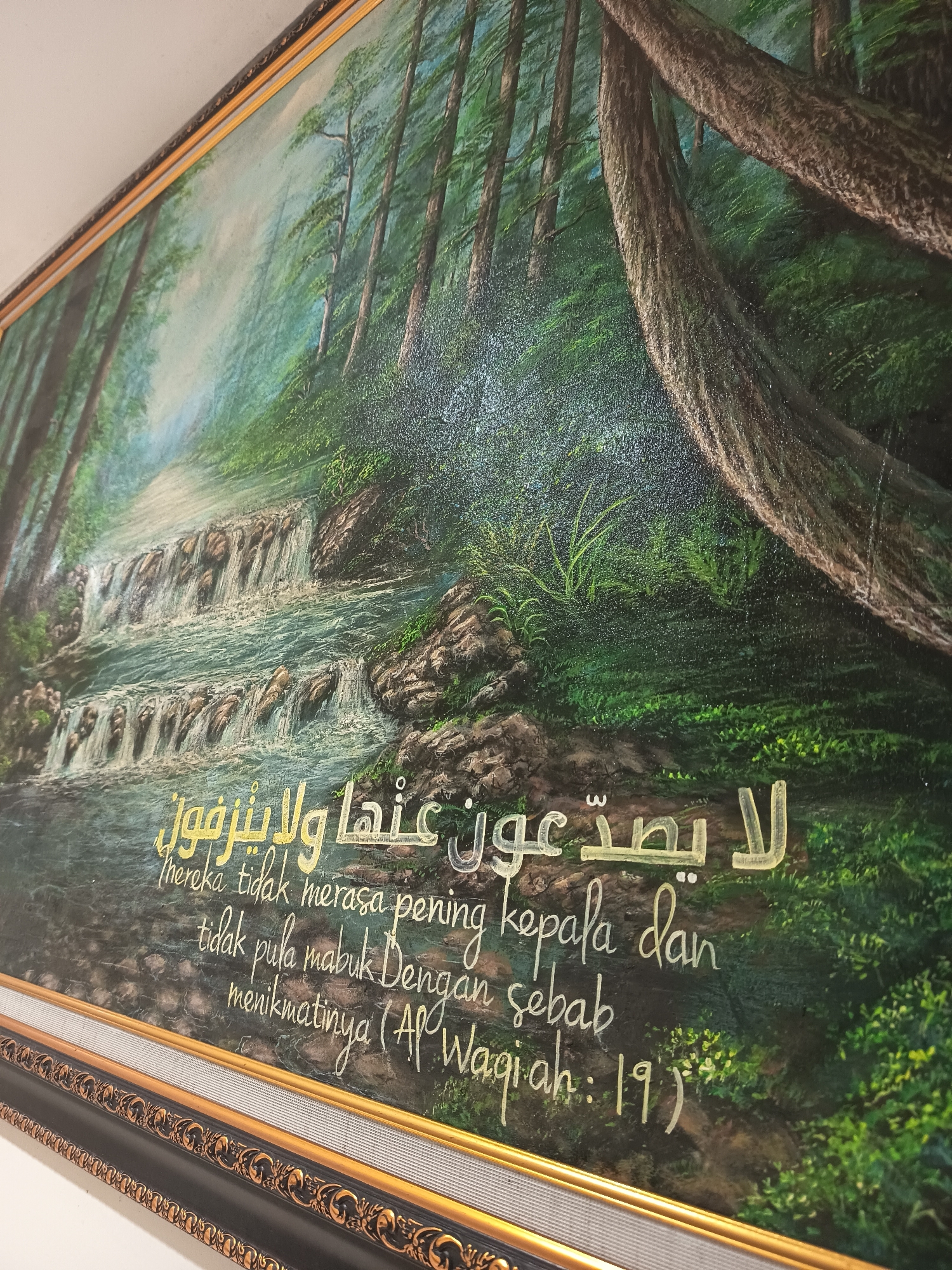 The image is a close-up, slightly angled photograph of a framed painting. The painting depicts a serene forest scene with a waterfall cascading down rocky terrain into a body of water. Lush green trees and foliage dominate the background, with thick tree trunks in the foreground. Overlaying the painting, in gold Arabic calligraphy and English text, is a verse that reads: "لا يصدعون عنها ولا ينزفون mereka tidak merasa pening kepala dan tidak pula mabuk Dengan menikmatinya (Al Waqiah: 19)". The frame is ornate, featuring a black base with gold detailing and a textured inner border. The context of Dapur, Indonesia, is not visually present in the image itself.
