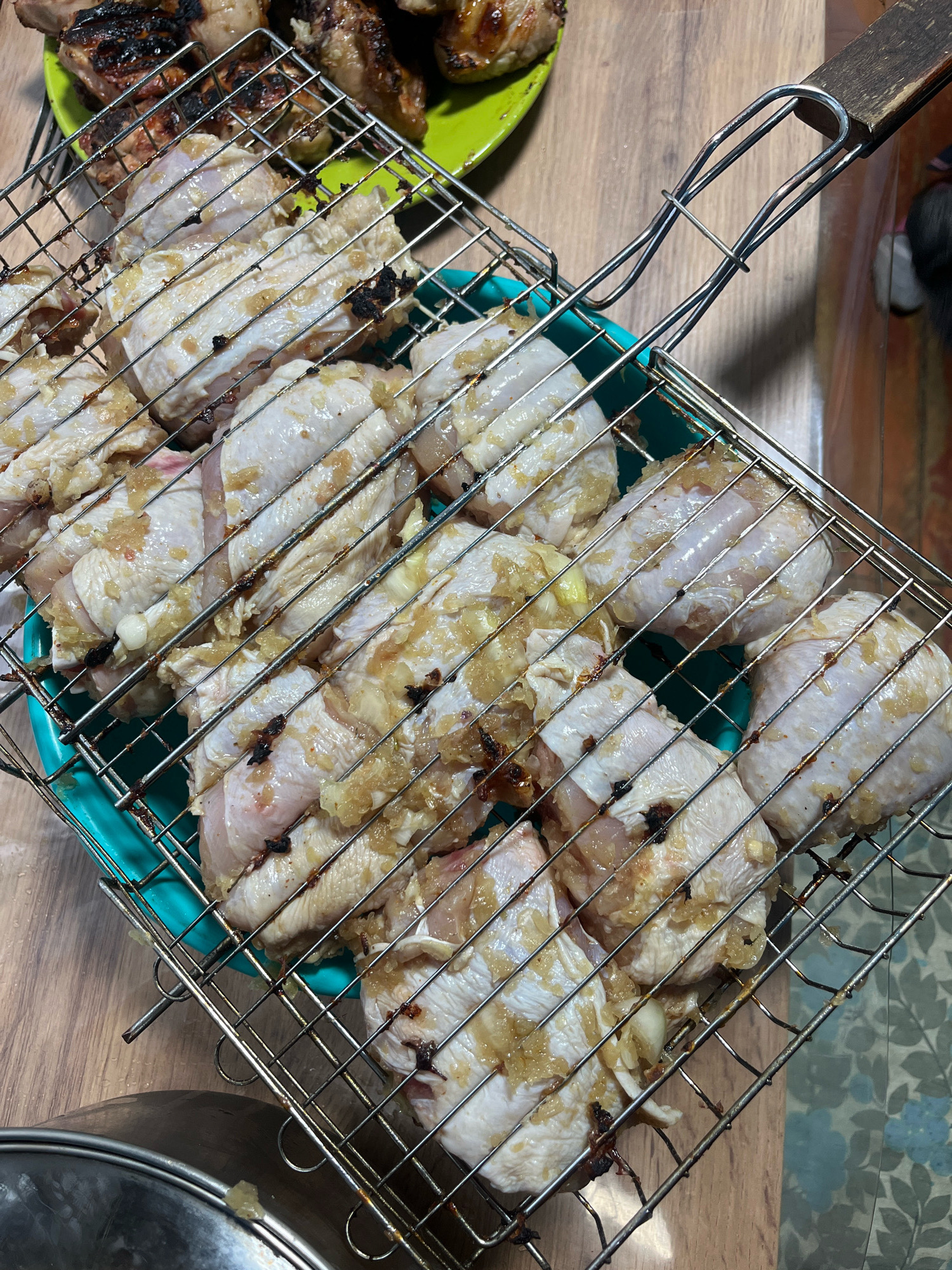The image shows a close-up, overhead view of chicken wings being prepared for grilling. The wings are arranged in a metal grill basket, which is placed on a wooden surface. Some of the chicken appears to have been seasoned with what looks like garlic and herbs, and there are some small charred spots indicating it may have been partially grilled already. In the background, a green plate with cooked chicken pieces is visible, as well as a glimpse of a patterned tablecloth. The overall scene suggests a home kitchen or outdoor cooking area. There is no indication of the specific location within Nizhny Tagil, Russia.