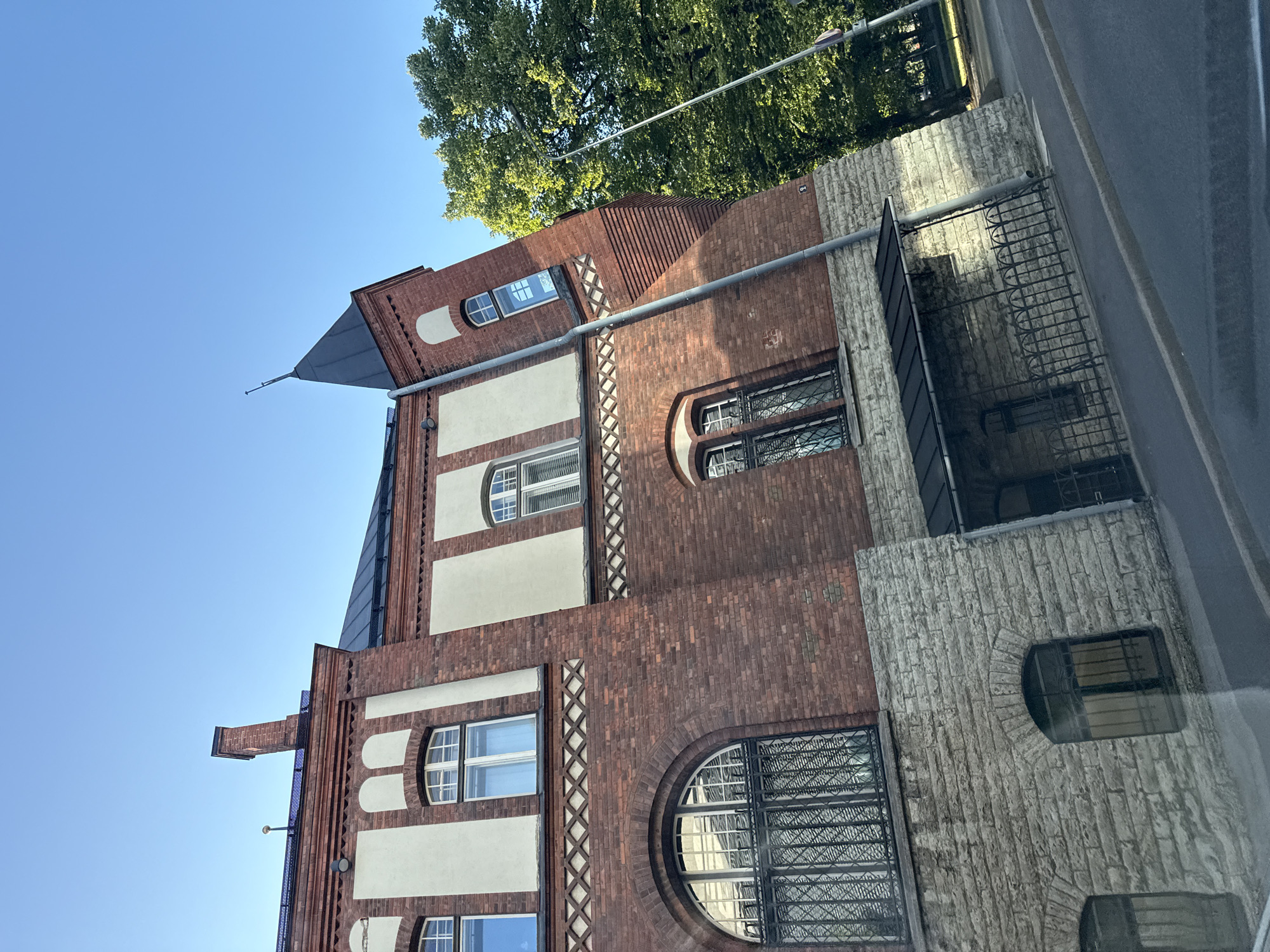 This is a photograph of the building located at Suur-Karja 18 in Tallinn, Estonia. The building is made of red brick and stone, with white plastered sections and arched windows. A pitched black roof with a spire caps the right side of the building, and a chimney is visible on the left. Green trees are present to the right of the building, and a street is in the foreground. The sky is clear and blue. There are no people or vehicles visible in the image.