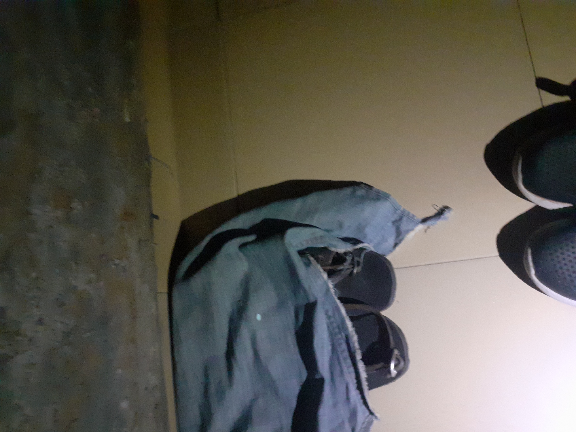 The image shows a low-angle view of a pair of dark-colored sandals partially covered by a piece of blue fabric, likely denim, lying on a tiled floor. In the foreground, the toe of a black athletic shoe is visible. The background consists of a textured, possibly concrete wall. The lighting suggests an indoor environment, possibly at night or in a dimly lit area, with a distinct light source casting shadows. The scene appears to be a mundane domestic setting, possibly Jalingo, Nigeria, but there is no direct evidence to confirm the location.