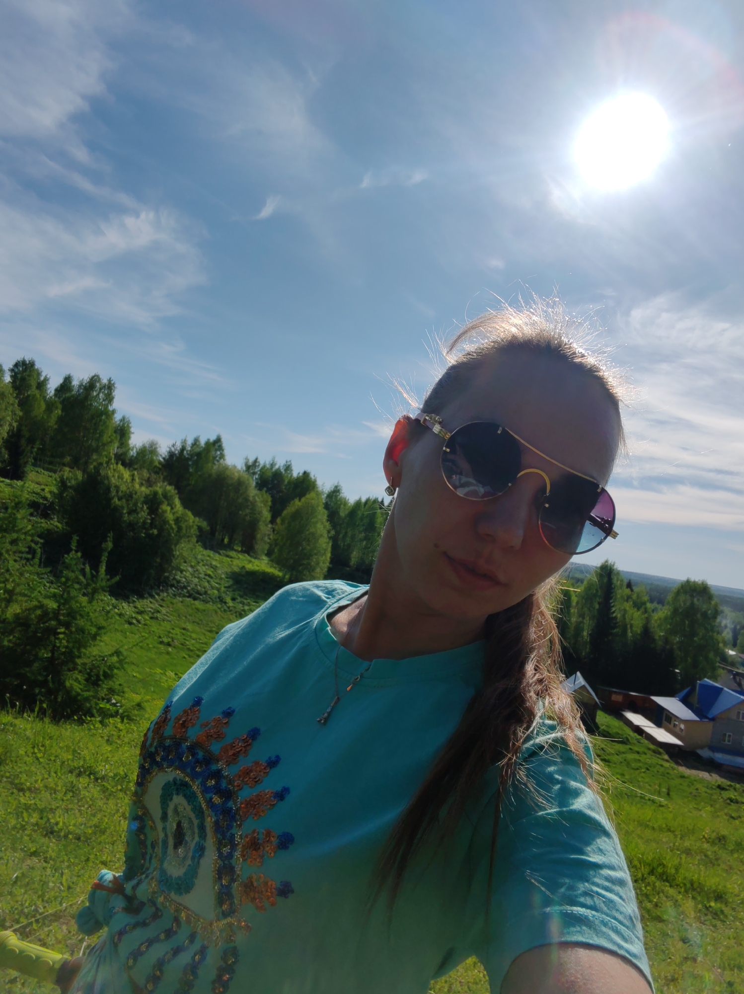 A woman with sunglasses is taking a selfie outdoors in Чухлэм, Russia. She is wearing a turquoise t-shirt with an eye design. A green hillside with trees is behind her. A bright sun shines in a blue sky with scattered clouds. Buildings are visible on the lower portion of the hillside.